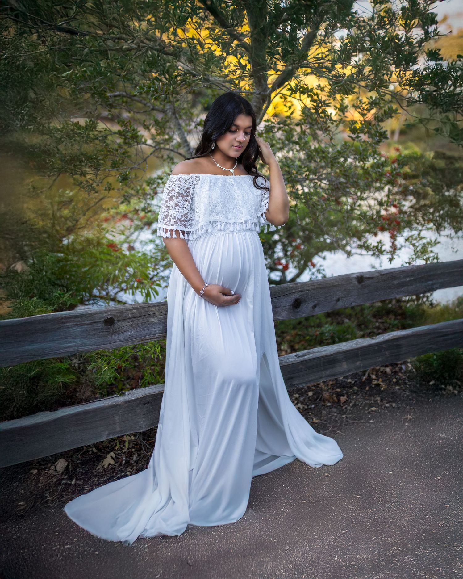 A pregnant woman in a flowing white lace dress poses by a wooden fence in a garden setting.