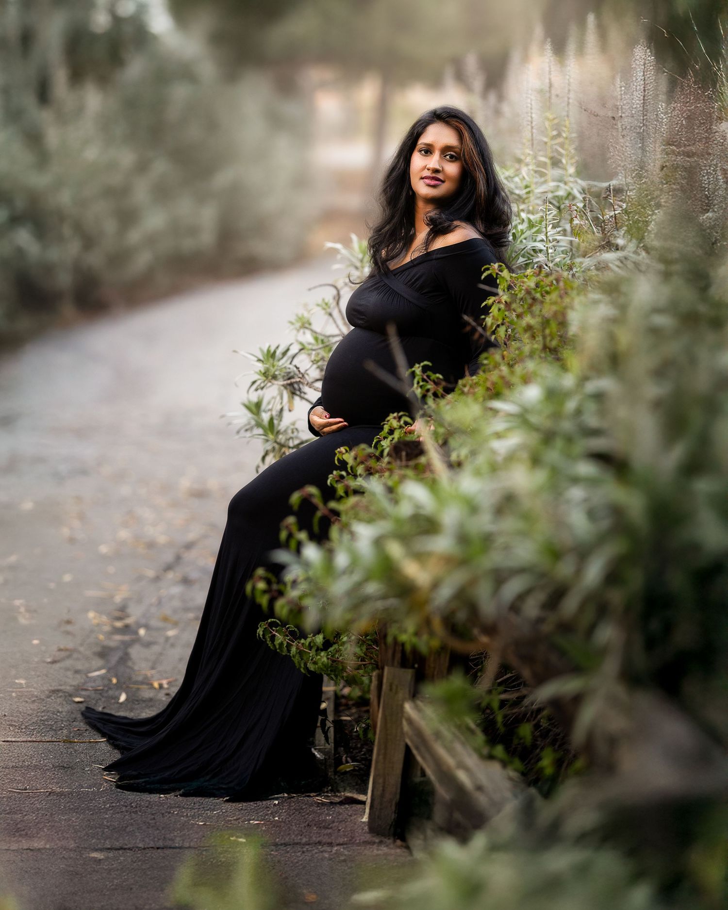 A person in a black dress poses gracefully on a bench along a wooded path during an outdoor photo session.
