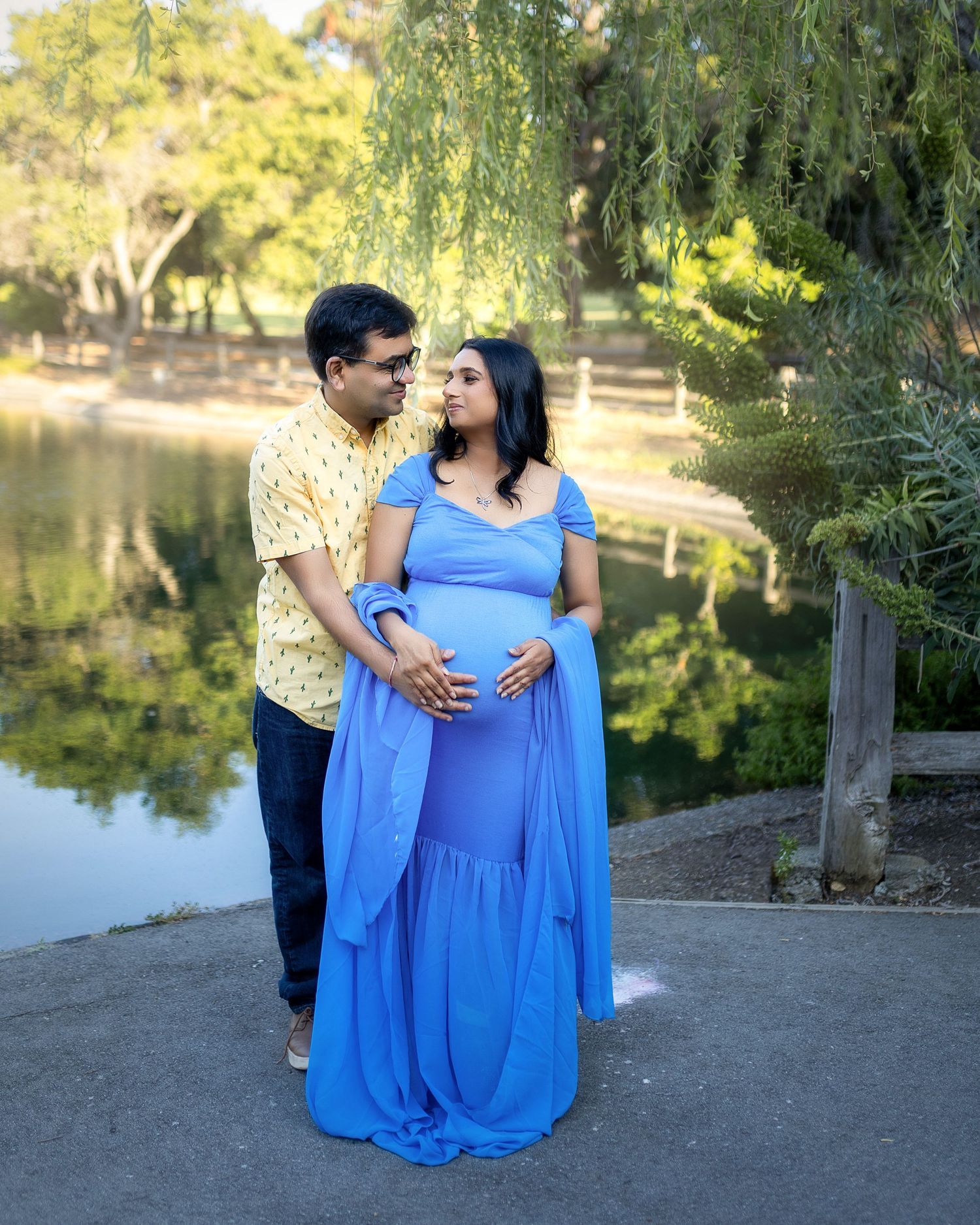 A couple in romantic maternity photo session by a serene pond with willow trees and natural lighting in the background.