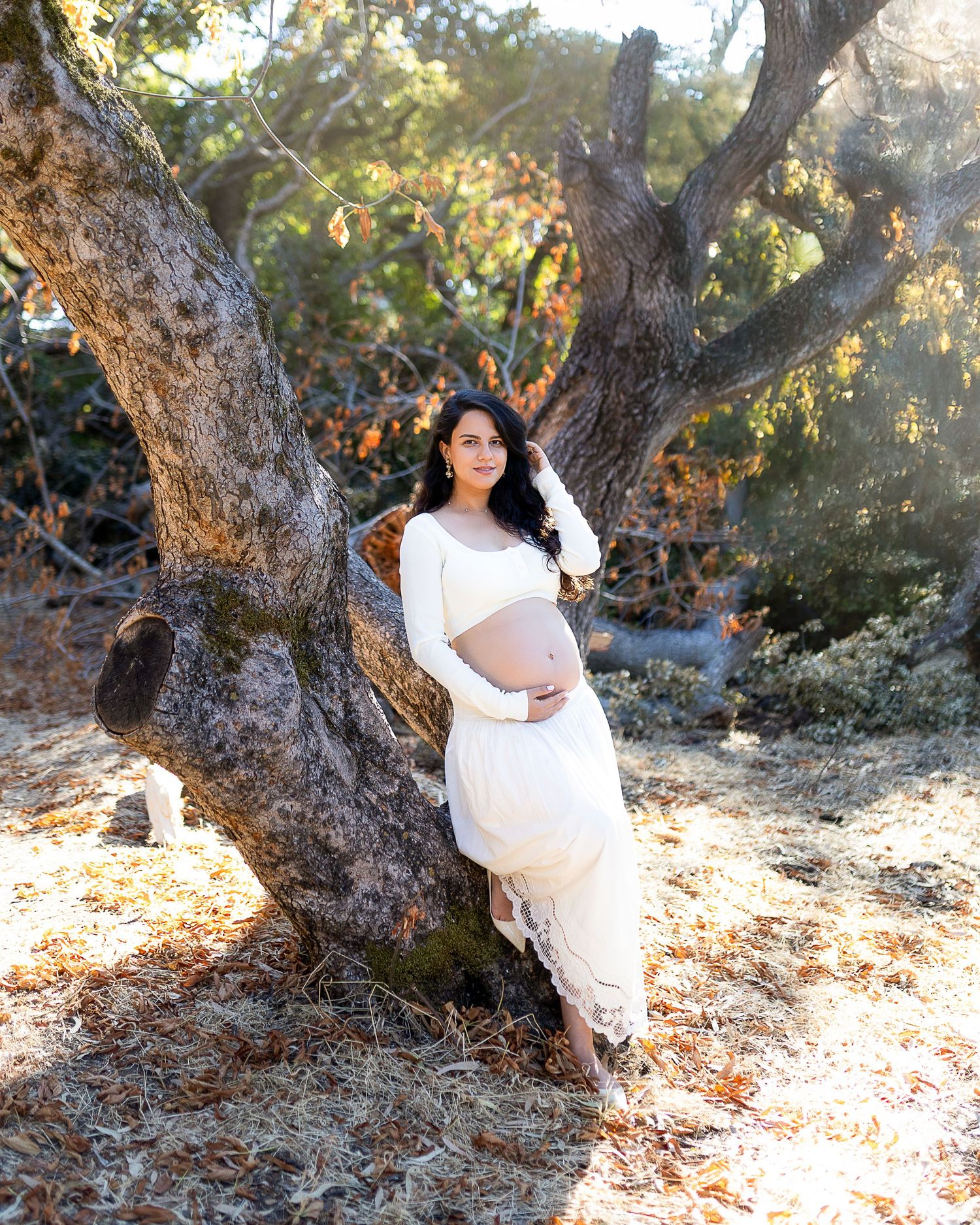 A figure in a flowing white dress poses gracefully among rustic trees in a natural outdoor setting.