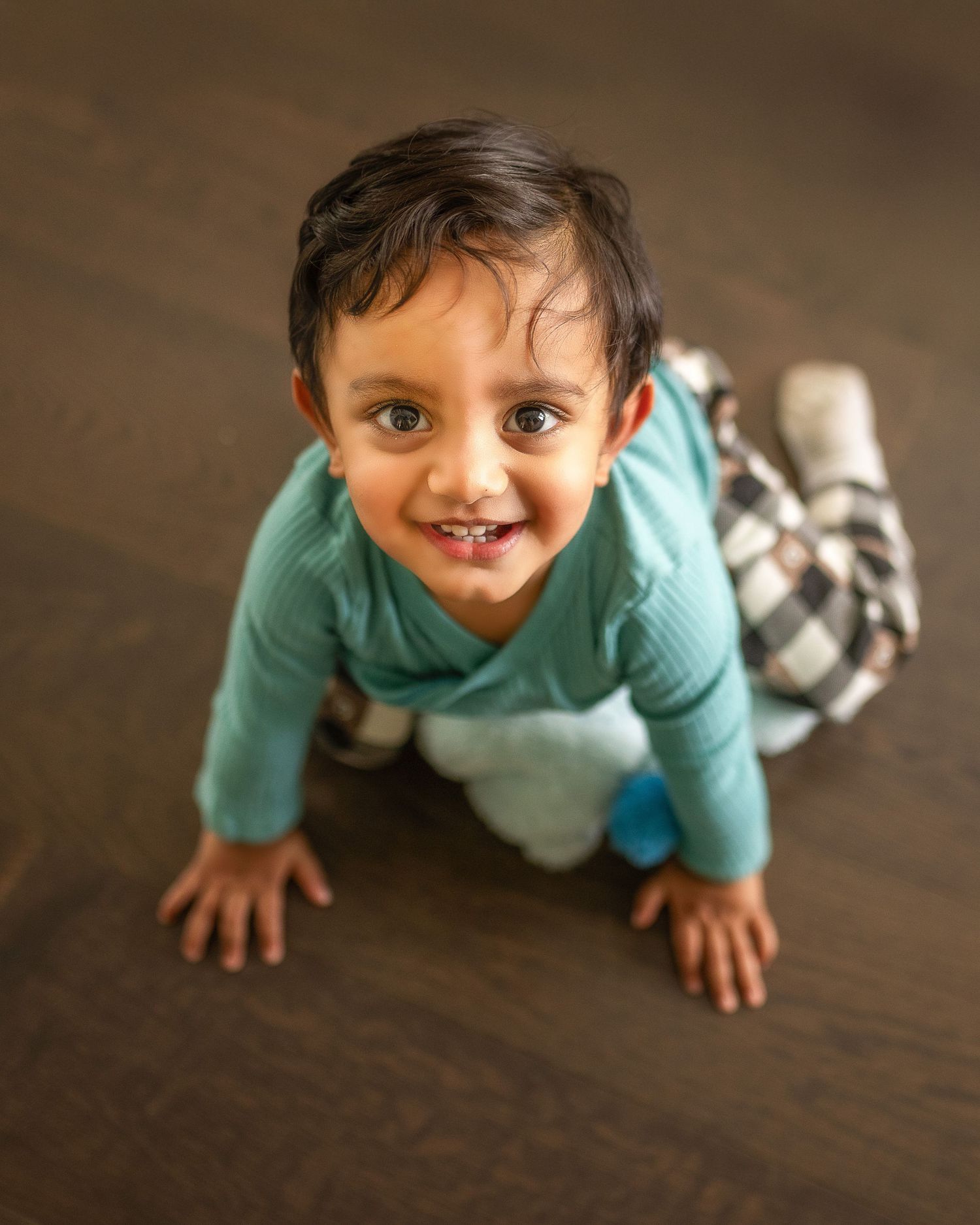 A small child in a mint green sweater and plaid pants crawls playfully on a hardwood floor.