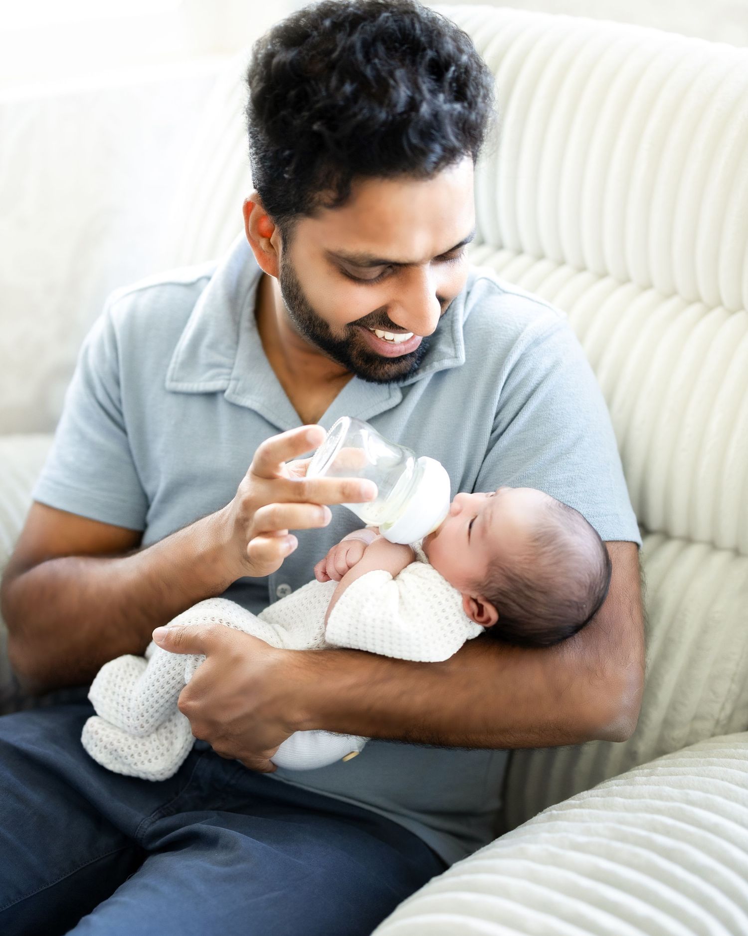 A parent sits on a white couch while bottle feeding their newborn baby and holding a small stuffed animal.