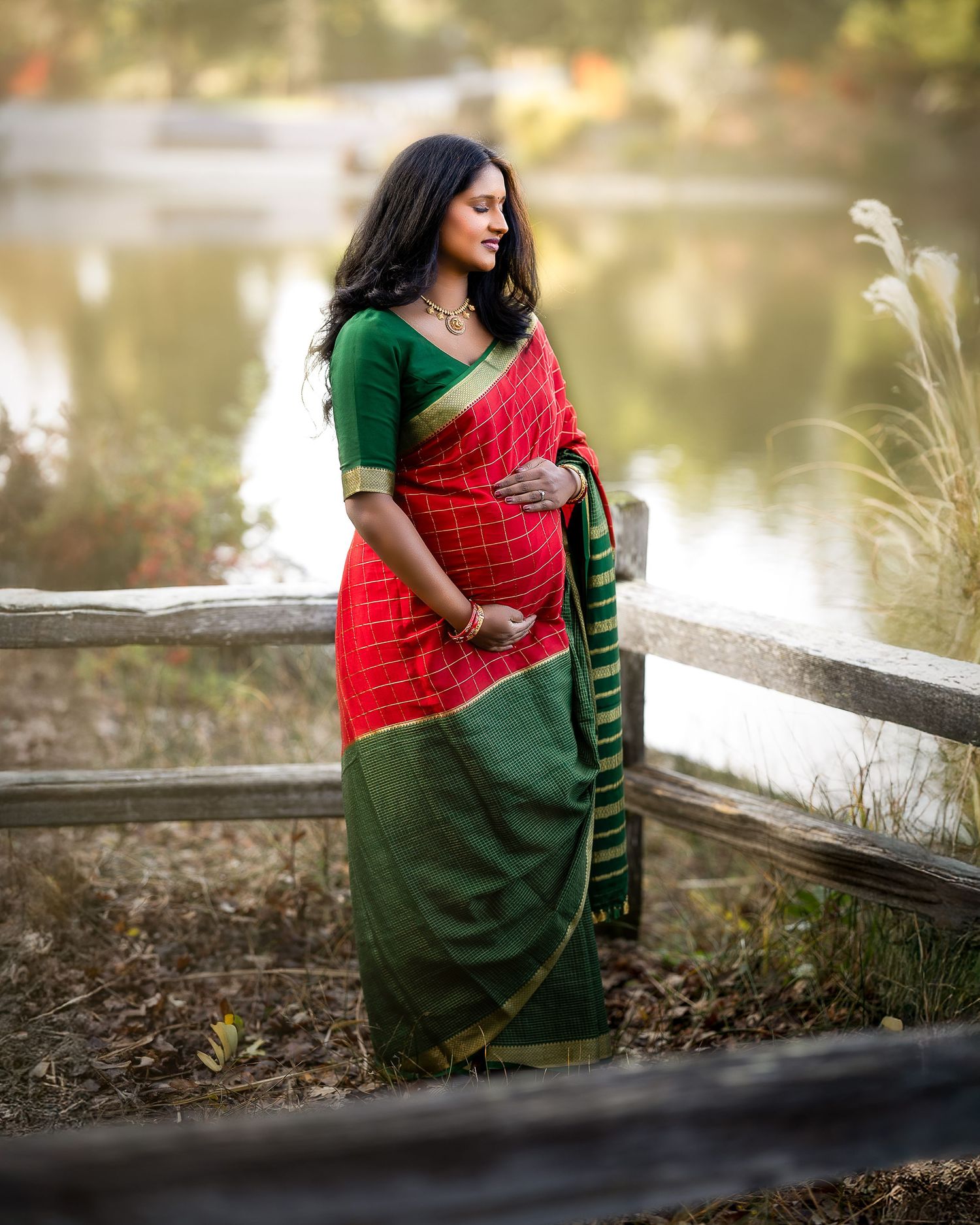 A person in a red and green saree sits gracefully by a serene lakeside during golden hour.