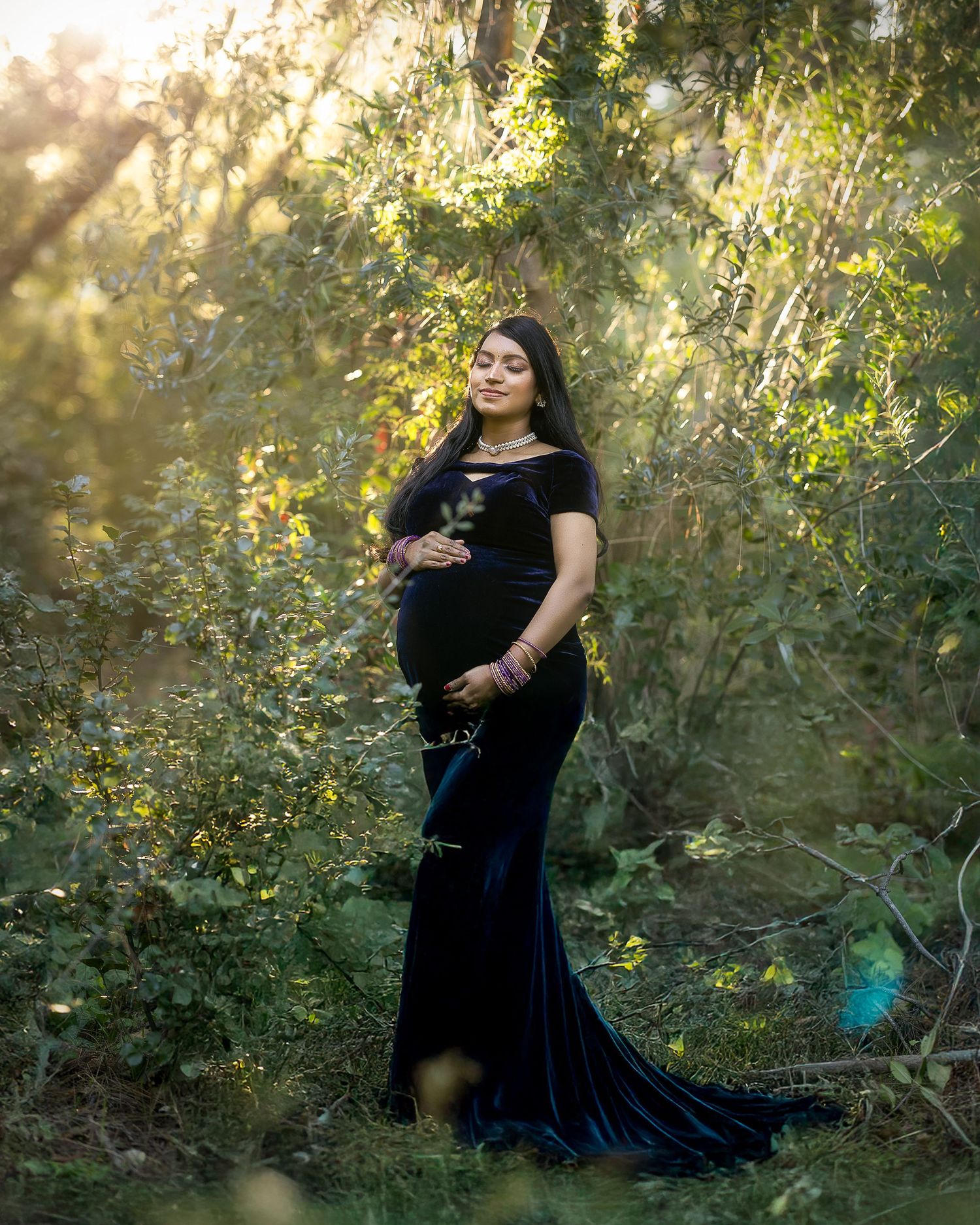 A pregnant figure in a black gown poses in a dreamy forest setting with sunlight filtering through the trees.