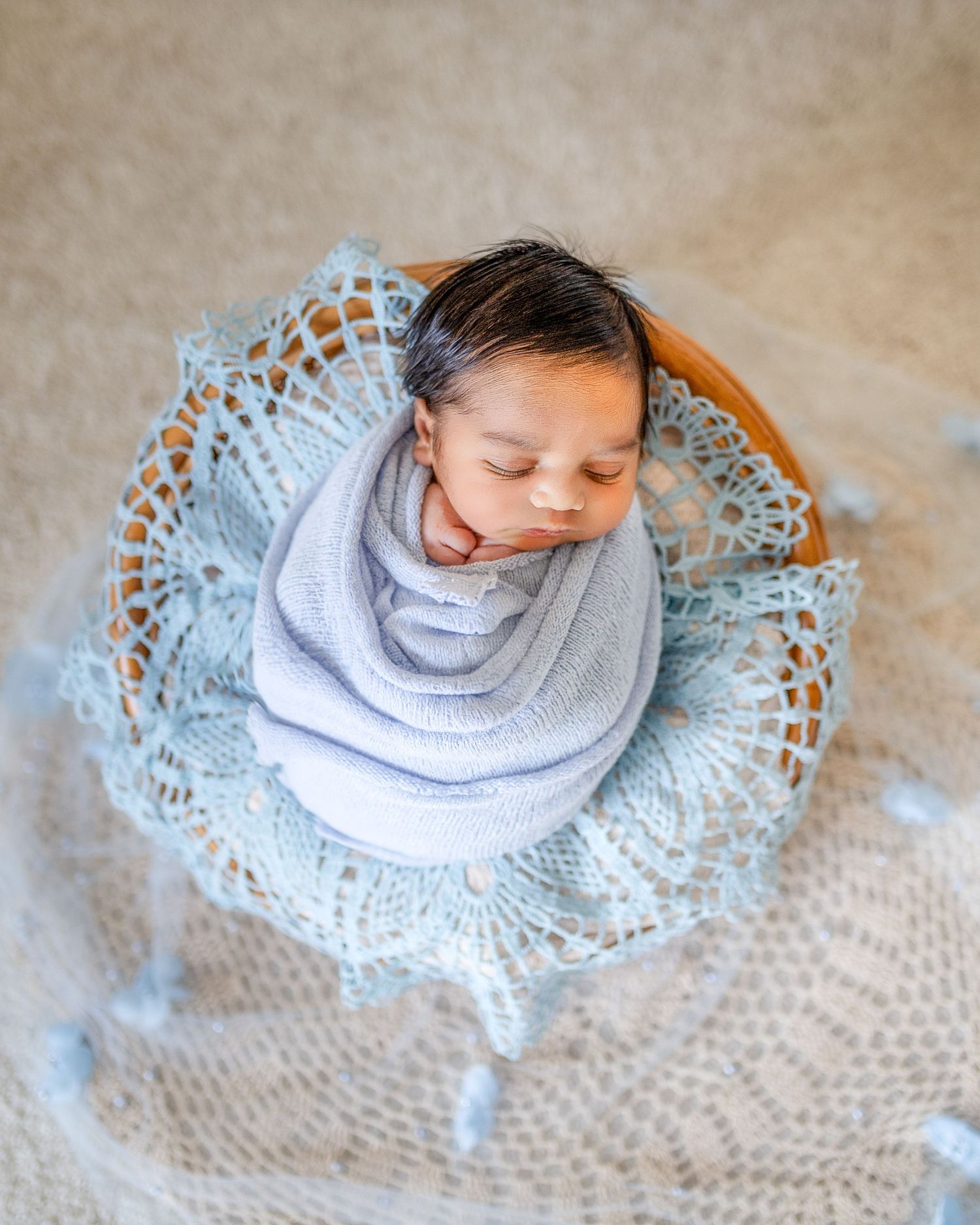 A child wearing a gray sweater lies on a circular blue crocheted doily on a beige carpet.