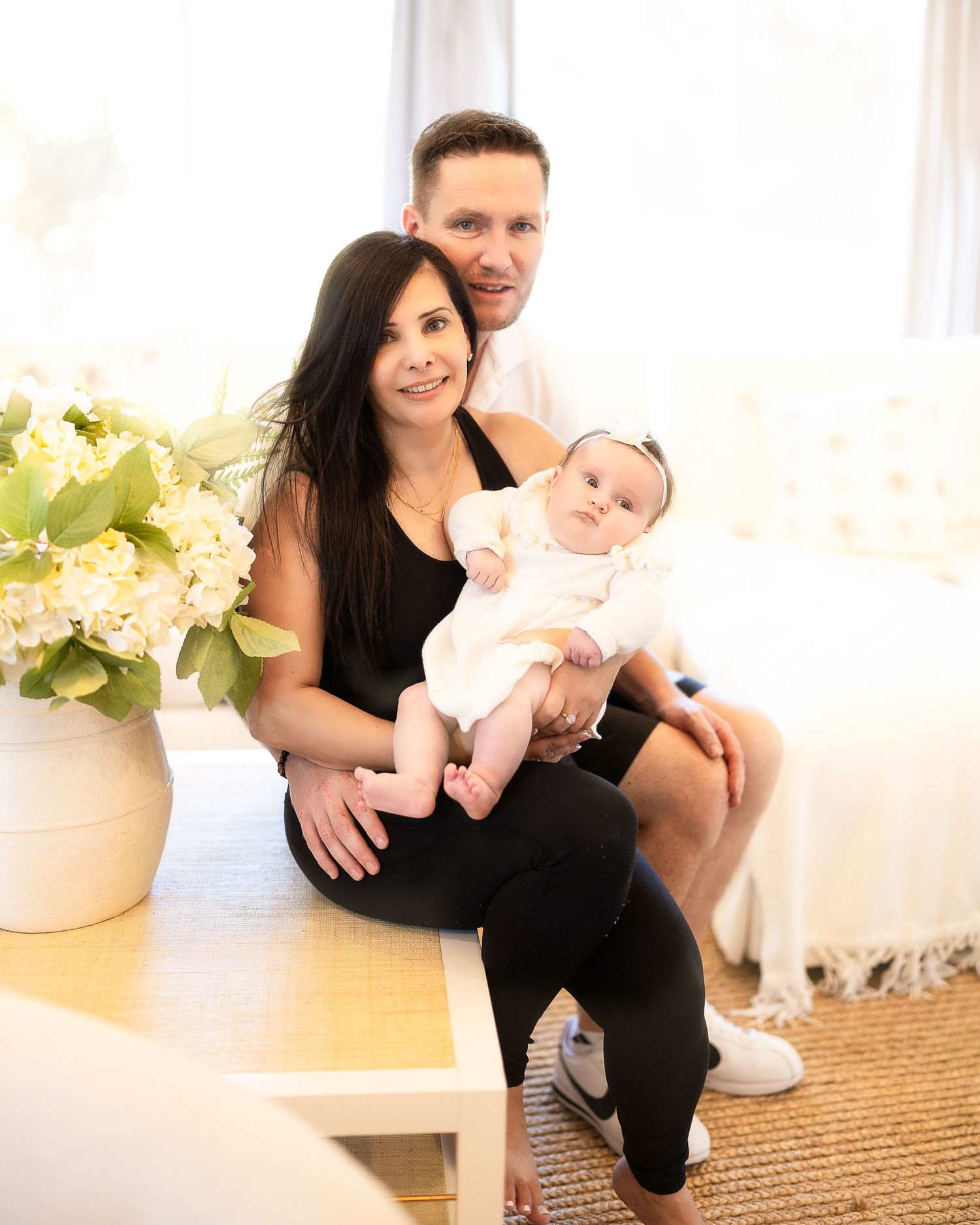 A family sits together on a bench with a small baby, next to a vase of white hydrangeas in a bright interior setting.