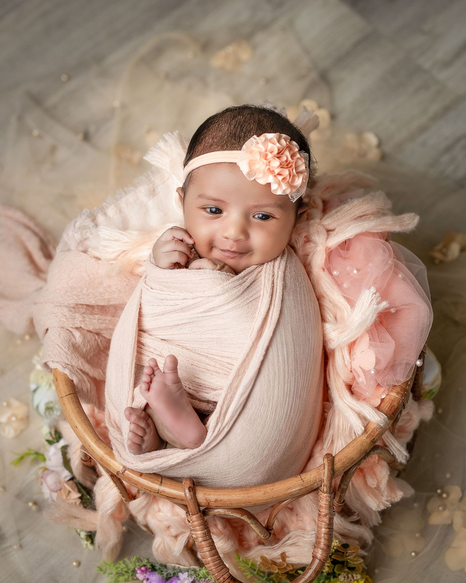 A newborn baby wrapped in pink fabric lies in a rustic wooden bowl surrounded by soft floral decorations.