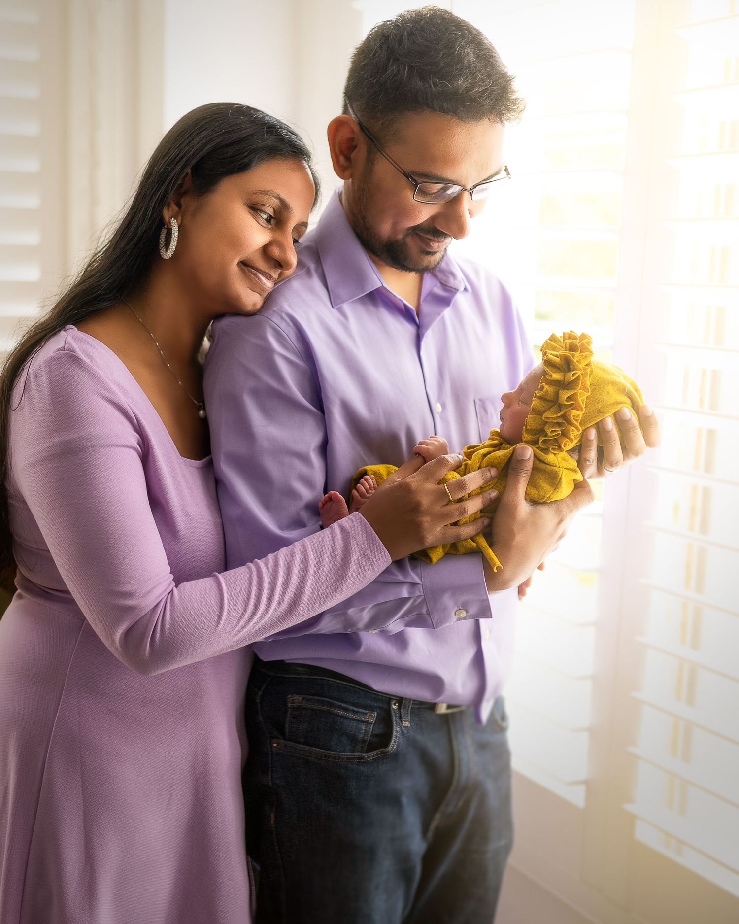 A couple in matching purple outfits share an intimate moment while holding a yellow teddy bear in a bright room.