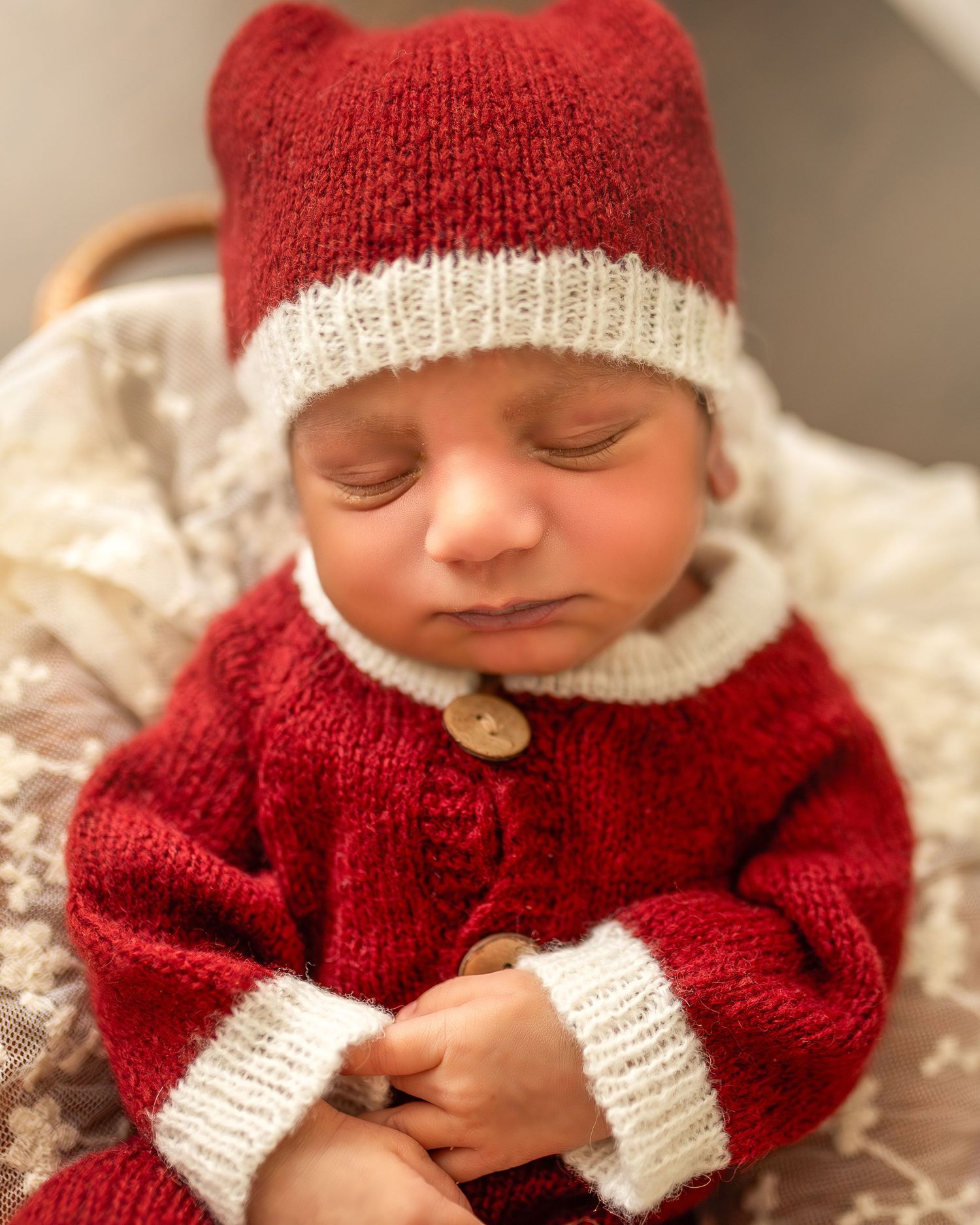 A baby wearing a red and white knitted sweater and hat lies peacefully on white bedding.