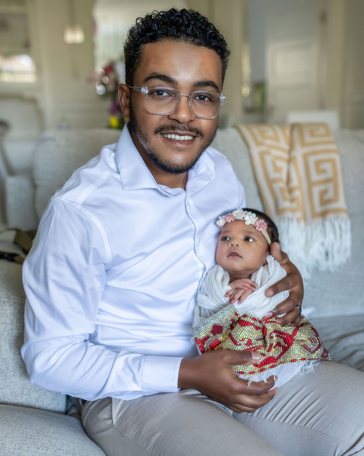 A person in a white shirt holds a small baby wearing traditional Indian clothing while sitting on a couch.
