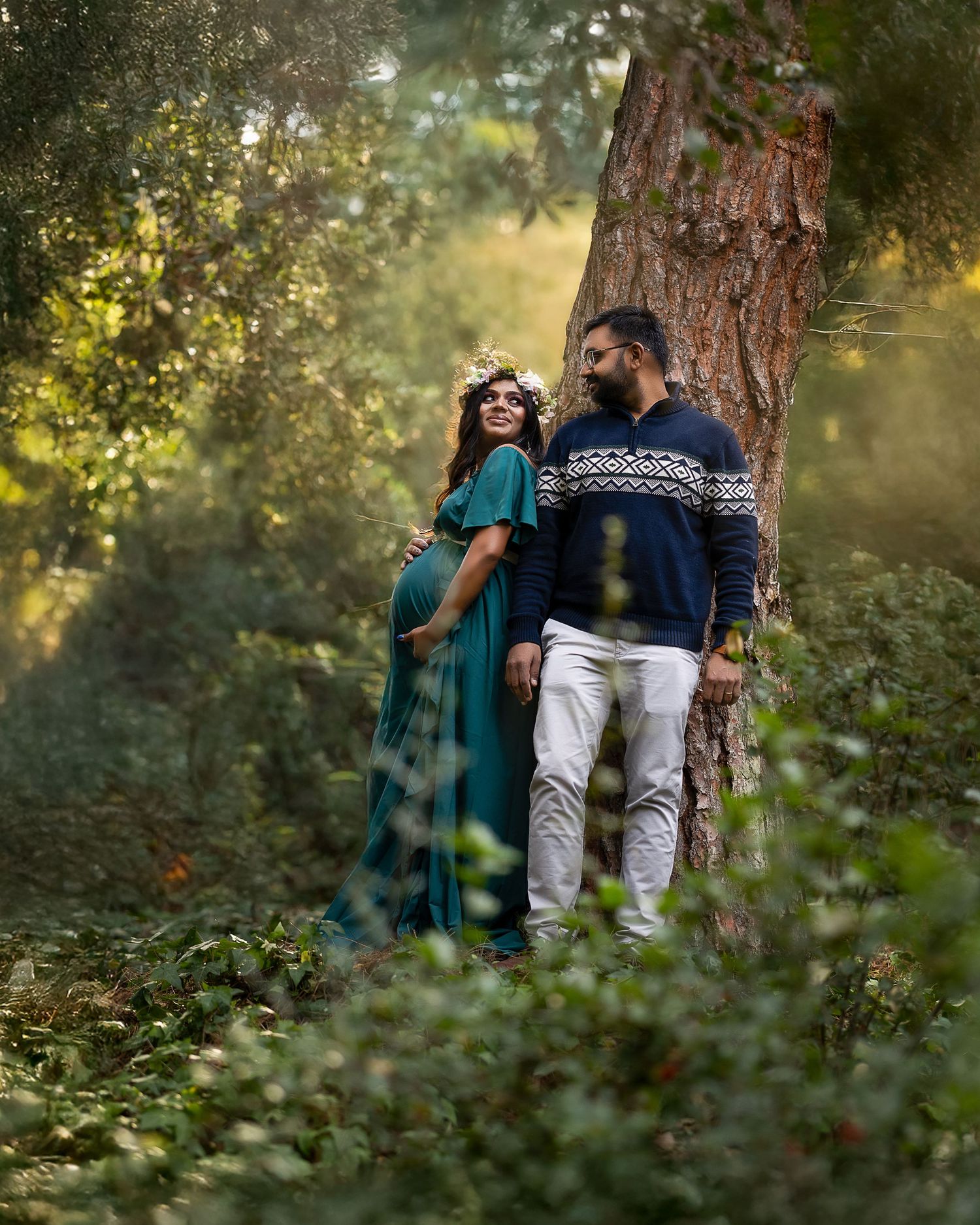 A couple in casual winter attire share a romantic moment against a tree in a dreamy forest setting.