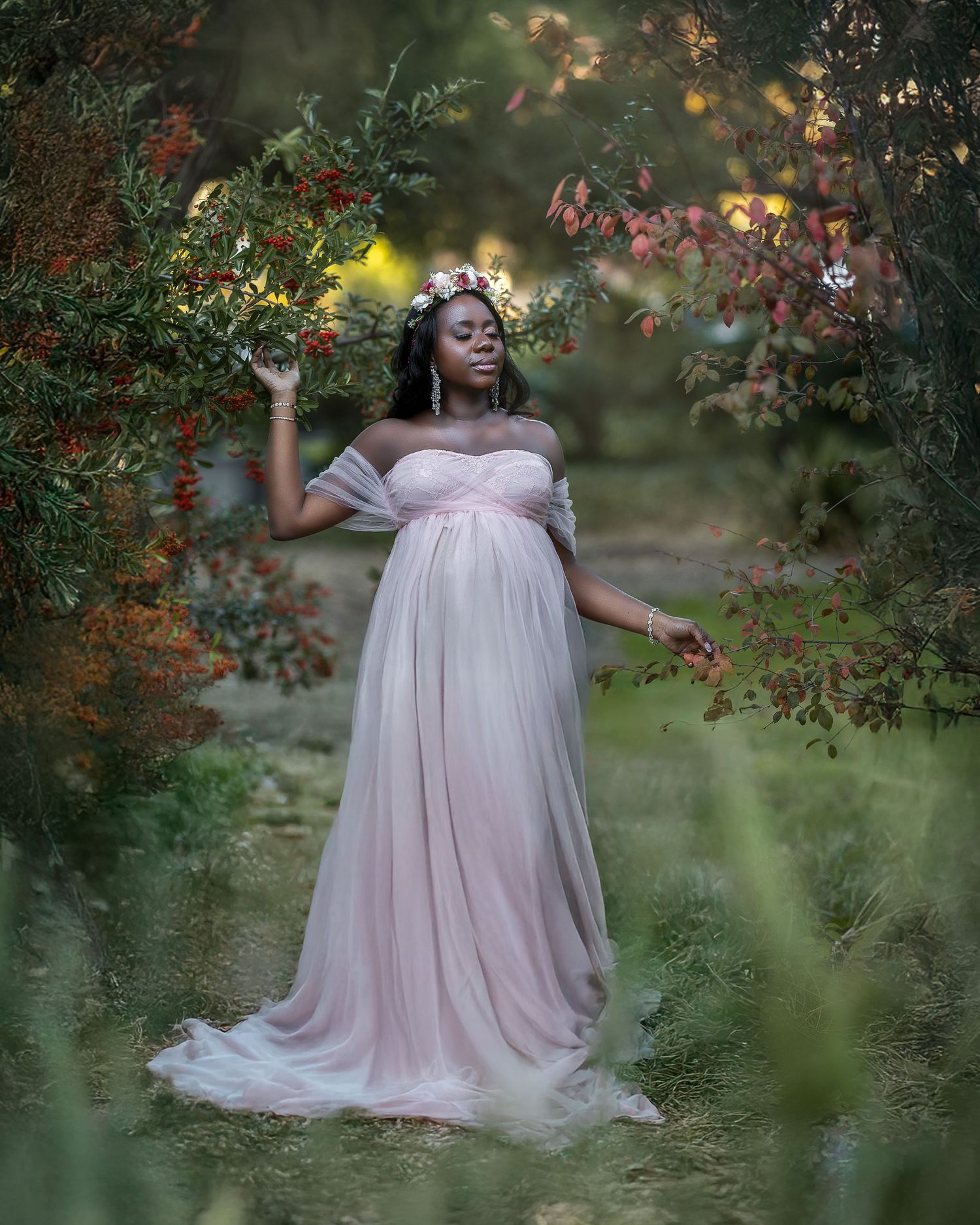 Ethereal outdoor maternity photoshoot featuring a flowing pink tulle gown against lush greenery and flowering branches.