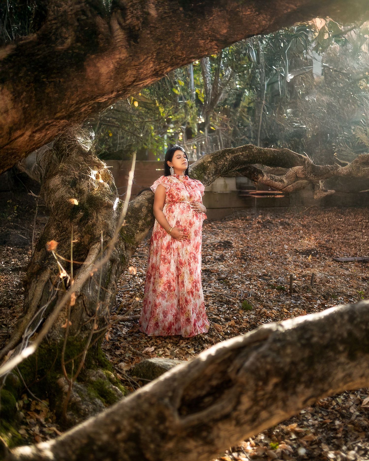 A person in a floral dress poses thoughtfully under a natural rock formation in a wooded setting during golden hour.