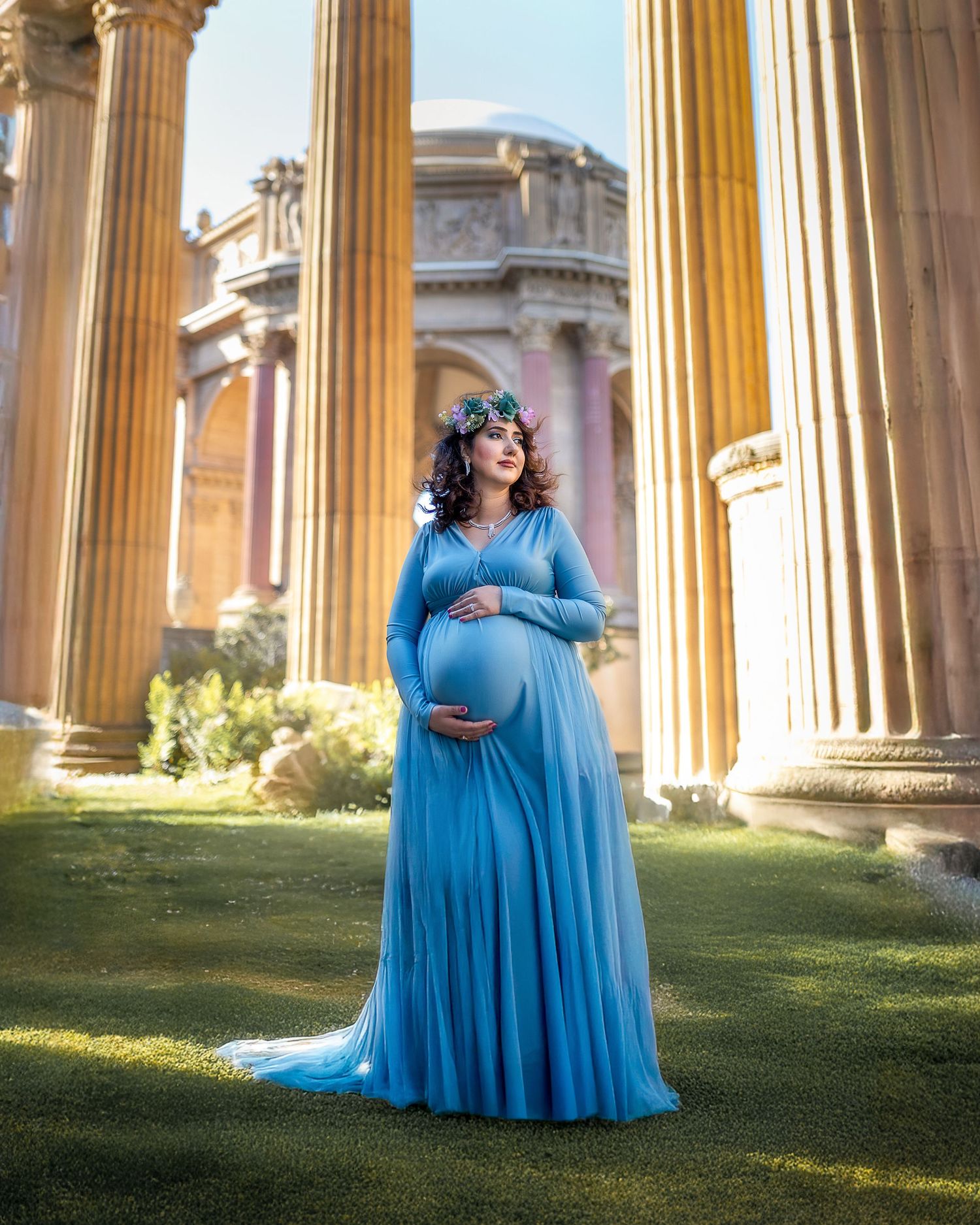 A flowing blue dress creates an ethereal silhouette against classical stone columns in a sunlit garden setting.