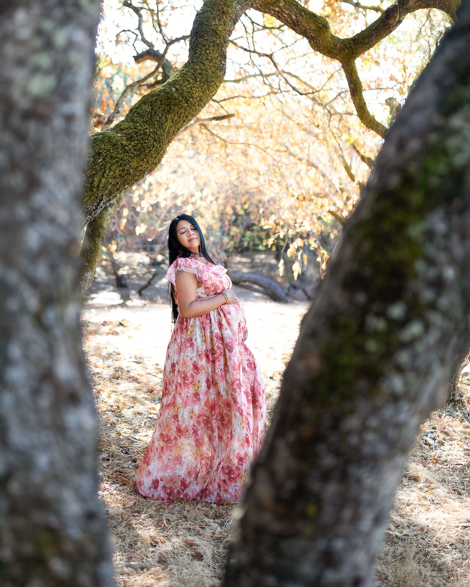 A woman in a floral pink dress poses among oak trees at golden hour in a dreamy outdoor setting.