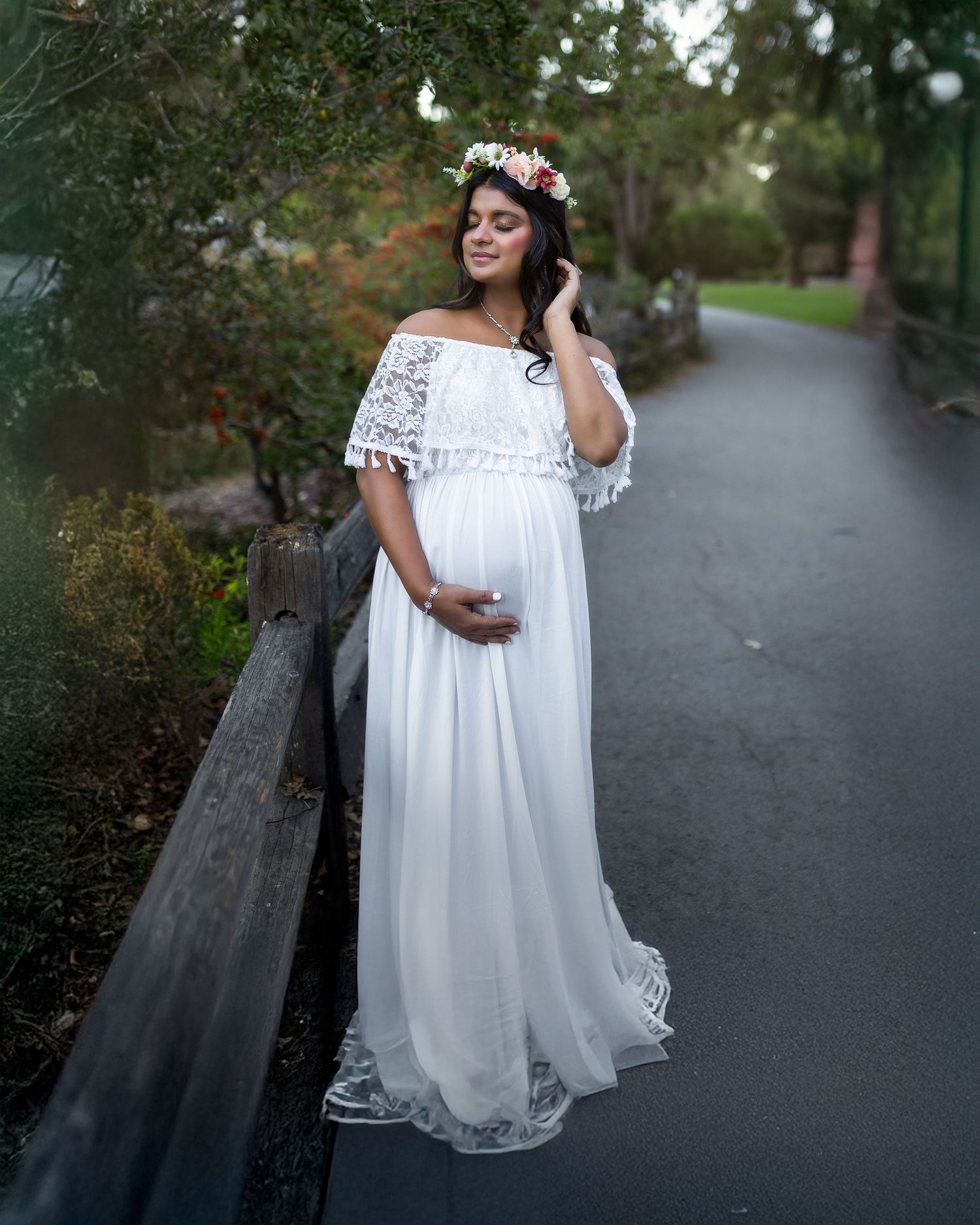 A pregnant woman in a flowing white lace maternity dress and flower crown stands on a garden path at sunset.