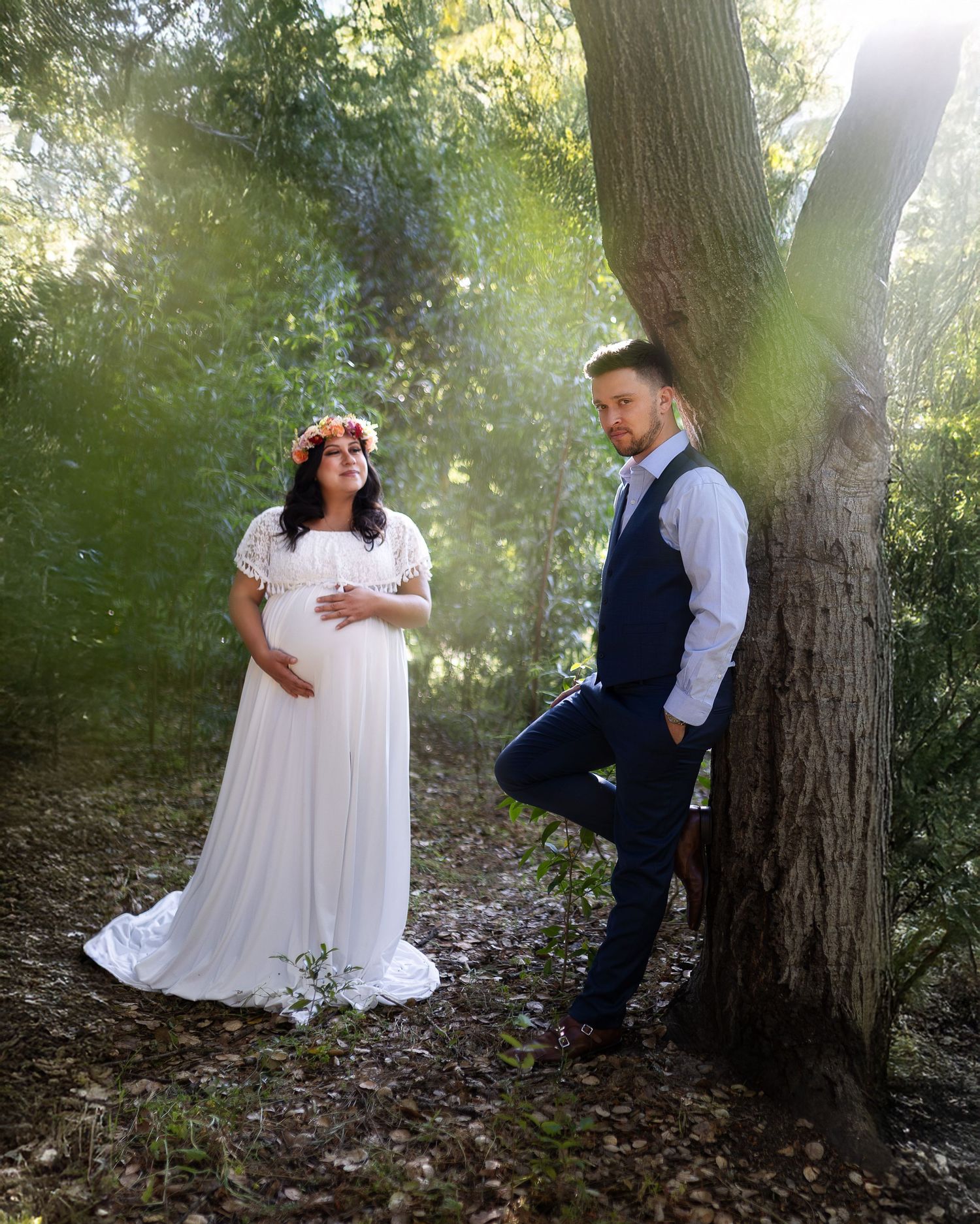 A romantic woodland photoshoot with a couple in white dress and formal wear posing by a large tree trunk in dappled sunlight.