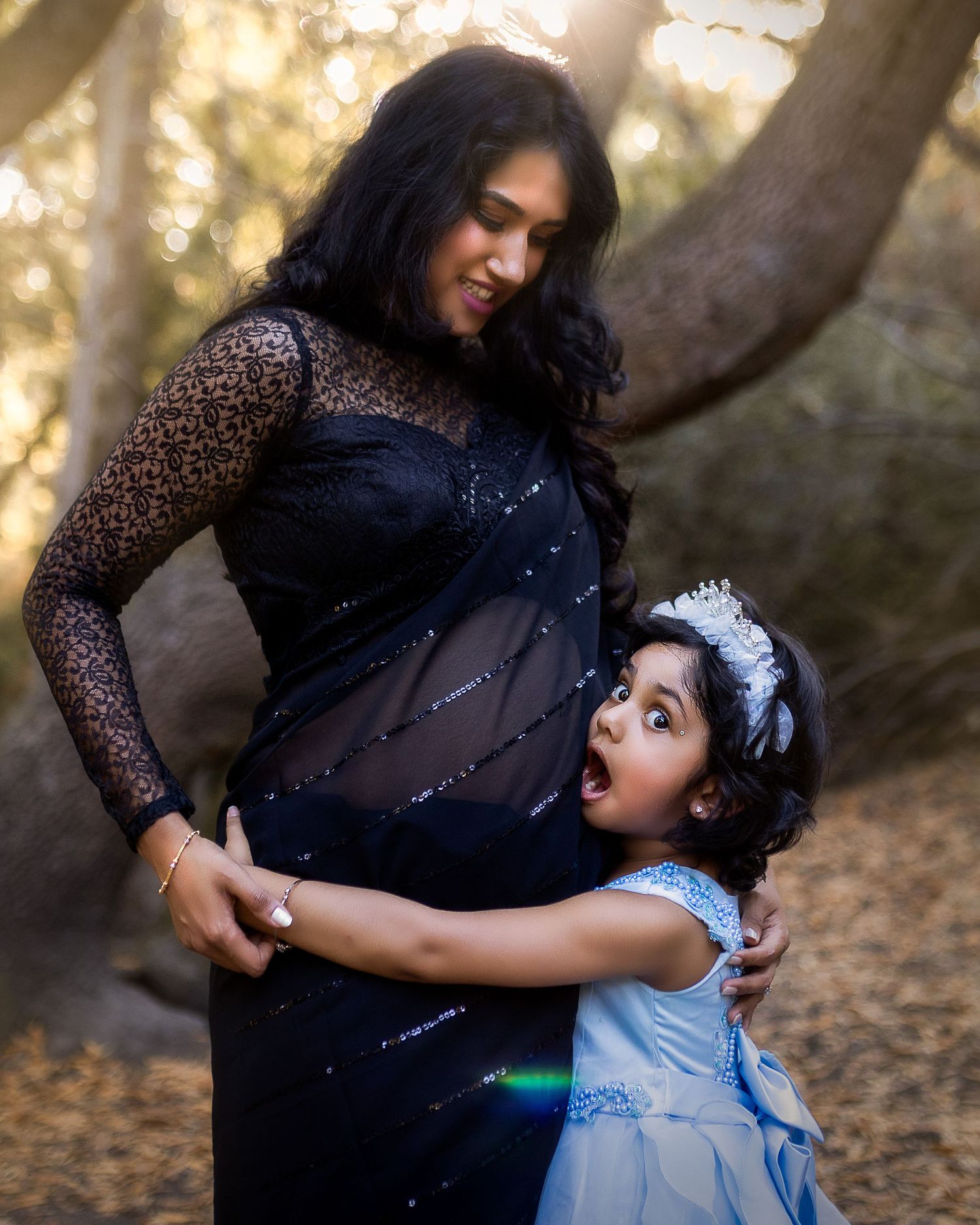A mother and daughter share a tender moment in a forest setting, wearing elegant dresses with sparkling details.