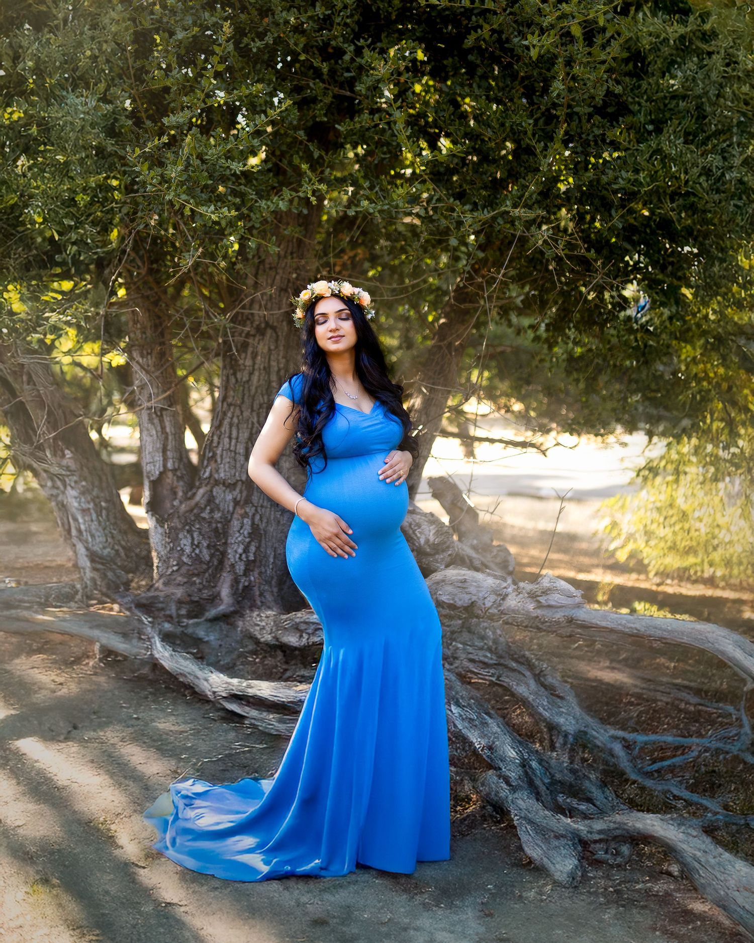 A person in a flowing royal blue dress poses gracefully near gnarled tree roots during a golden sunset photoshoot.