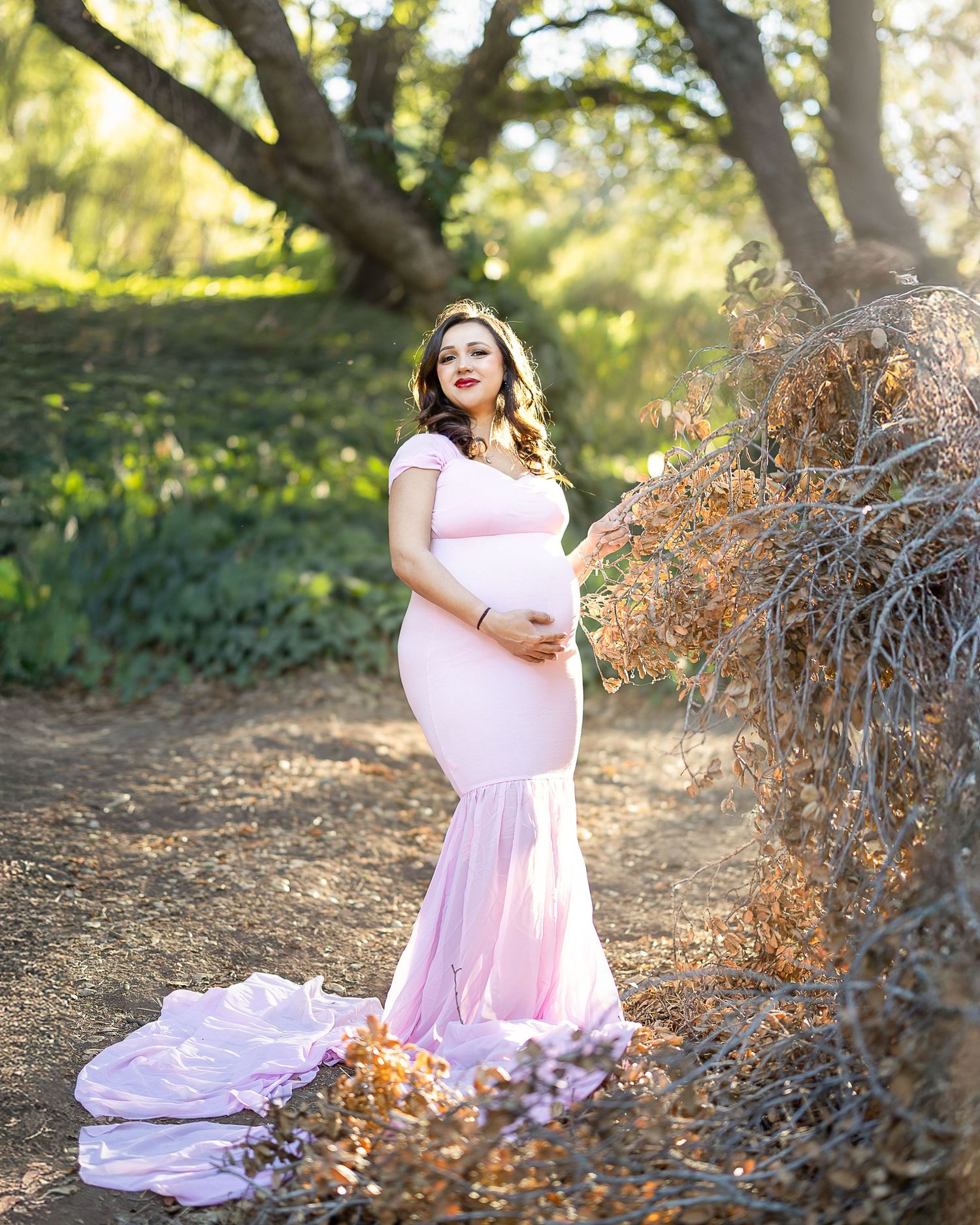 Expectant mother in flowing pink gown poses gracefully in a sunlit garden setting with trees and dried foliage.