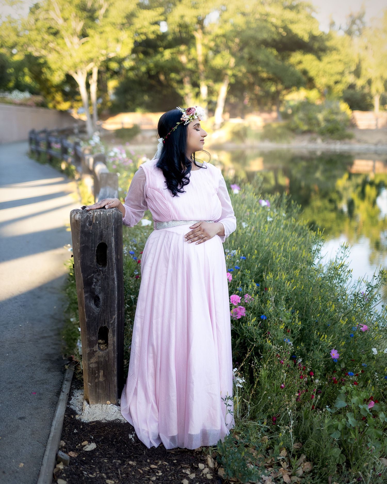 A person in a flowing pink dress stands among wildflowers along a lakeside path at sunset.