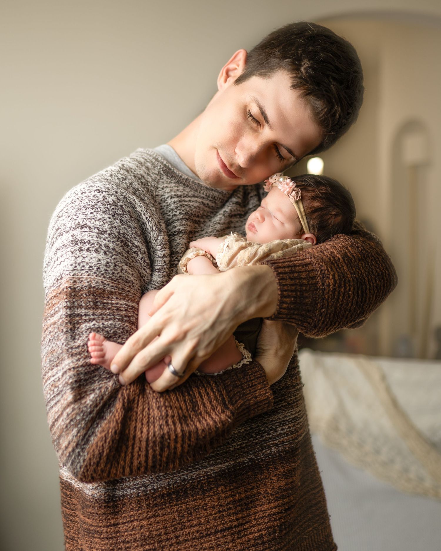 A young parent in a brown knit sweater tenderly holds a sleeping newborn baby close to their chest.