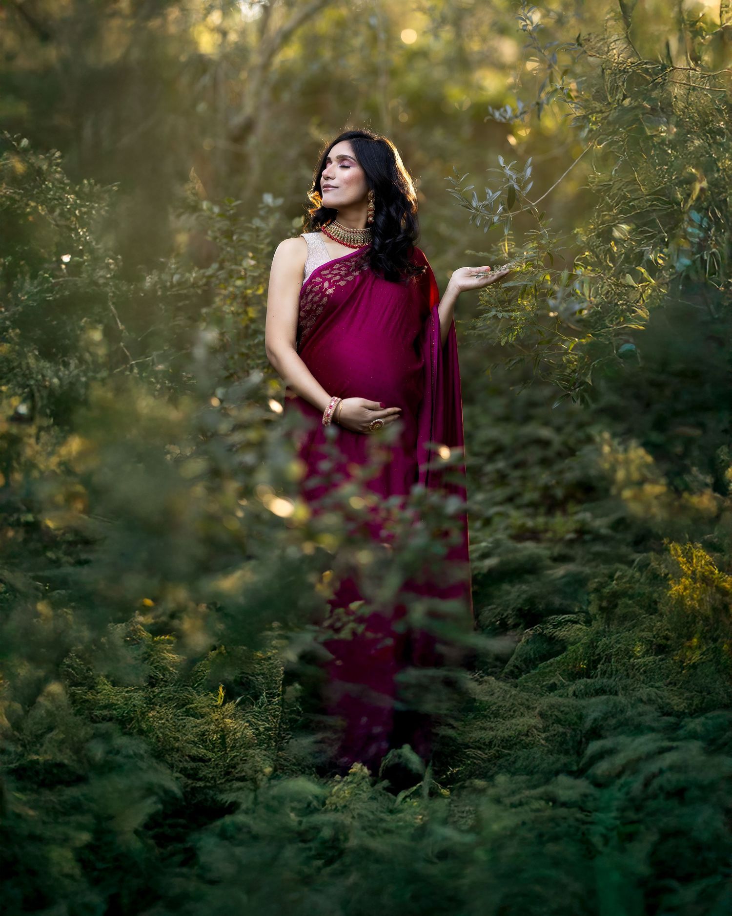 A figure in a flowing burgundy dress poses gracefully among lush foliage during golden hour.
