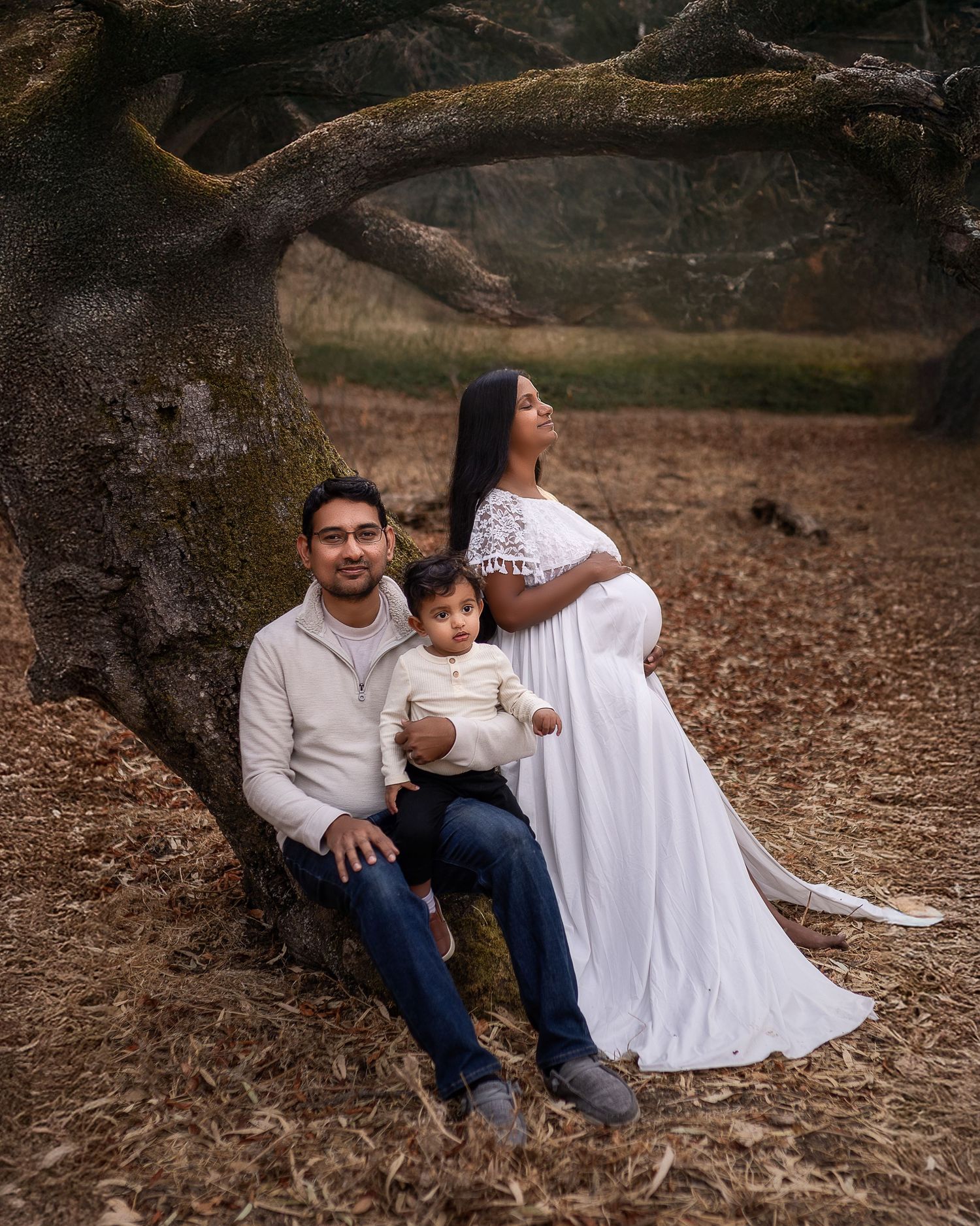 Family relaxes together on fallen leaves beneath twisted tree branches in a natural outdoor setting.