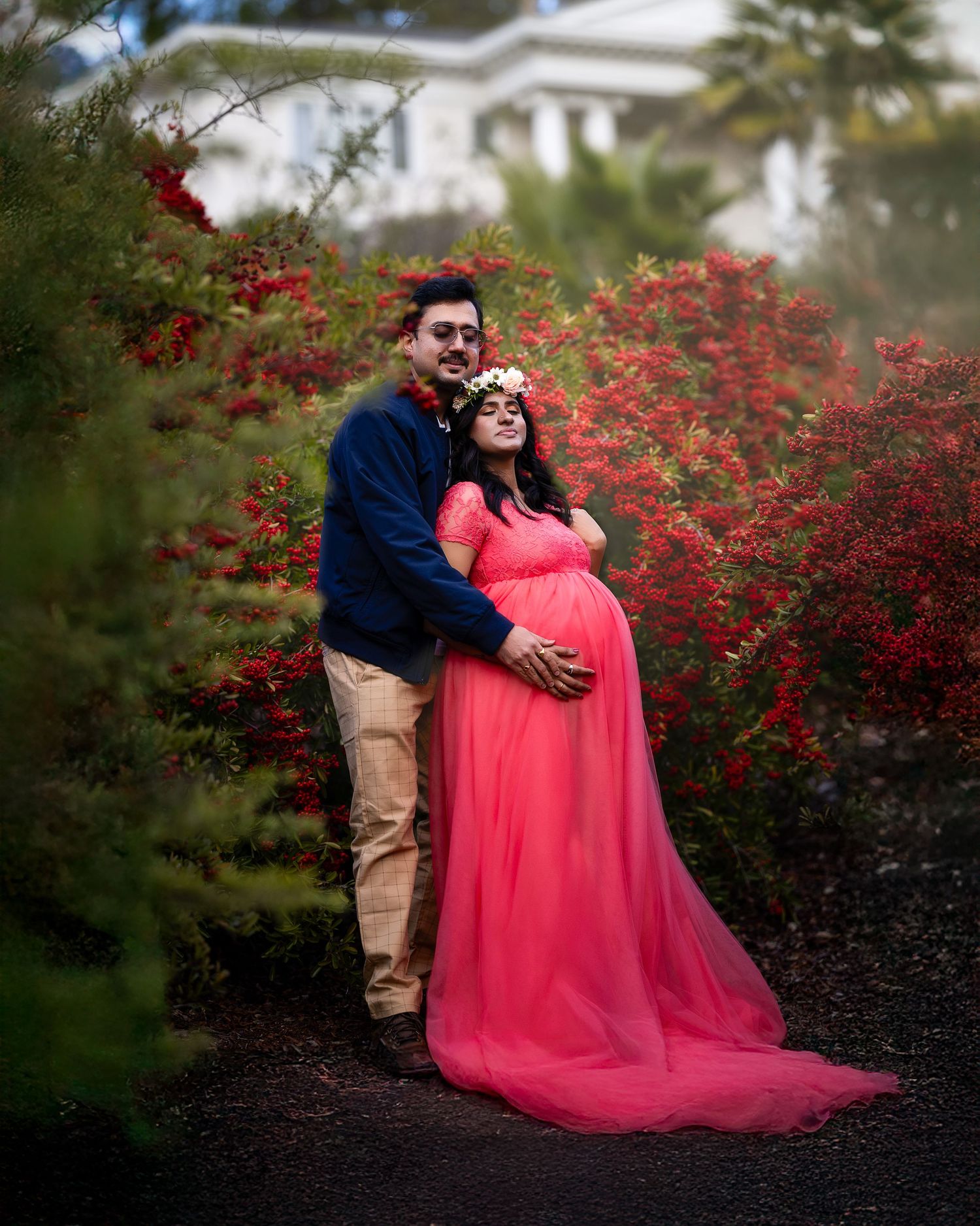 A couple poses together against vibrant red flowering bushes during a romantic outdoor maternity photoshoot.