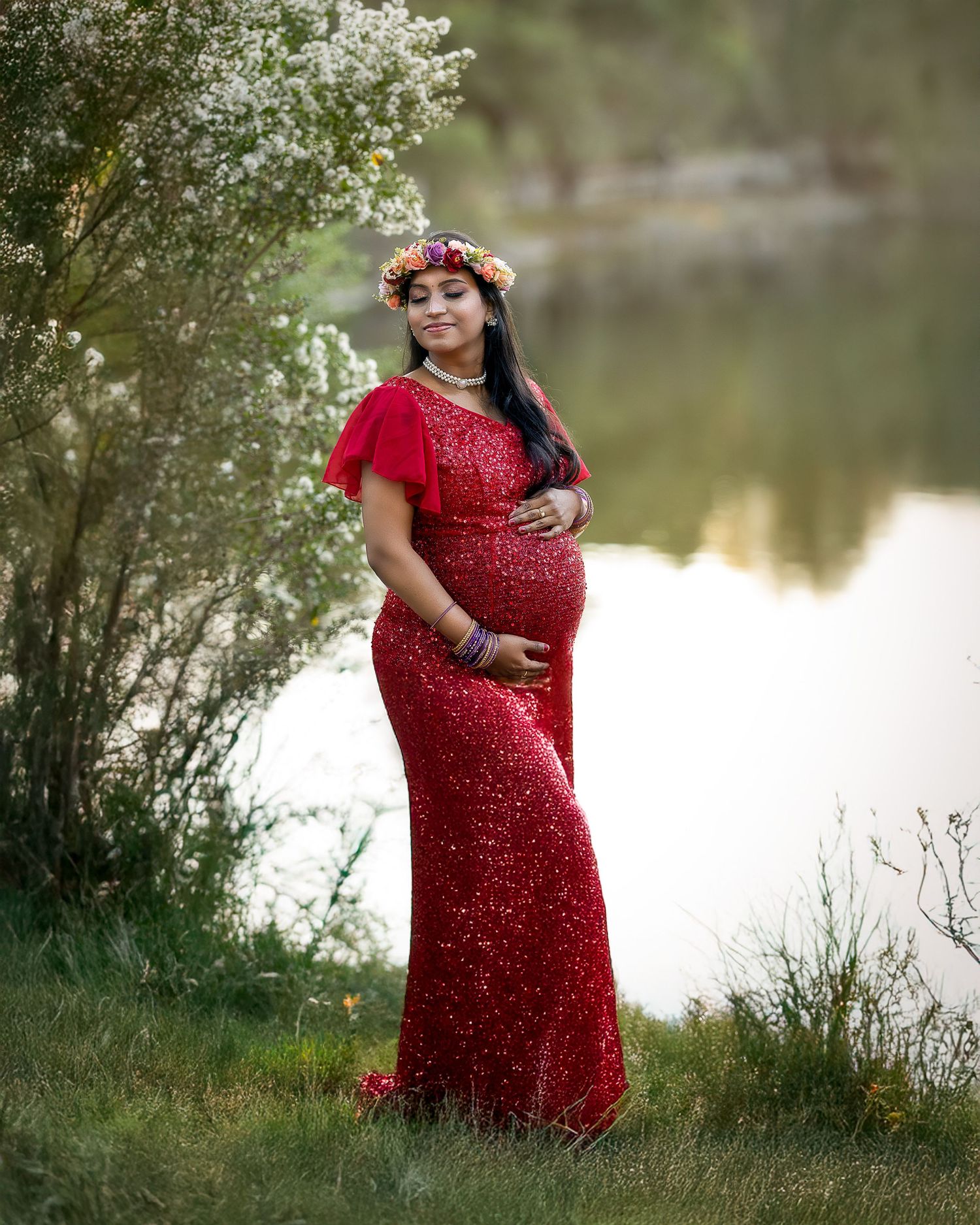 A pregnant woman in a sparkling red gown poses gracefully by a serene lake surrounded by lush greenery.
