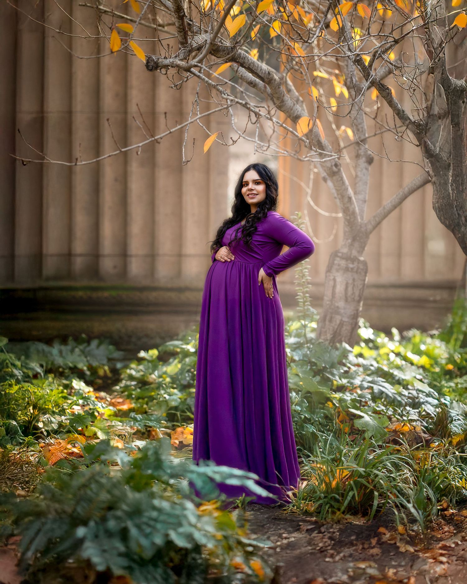 A figure in a flowing purple maxi dress poses among autumn foliage against a columned stone wall in soft lighting.