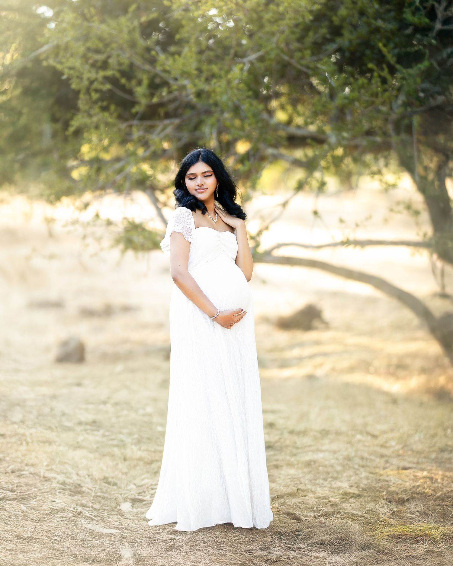 A person in a flowing white dress poses gracefully in a sun-dappled outdoor setting with trees in the background.