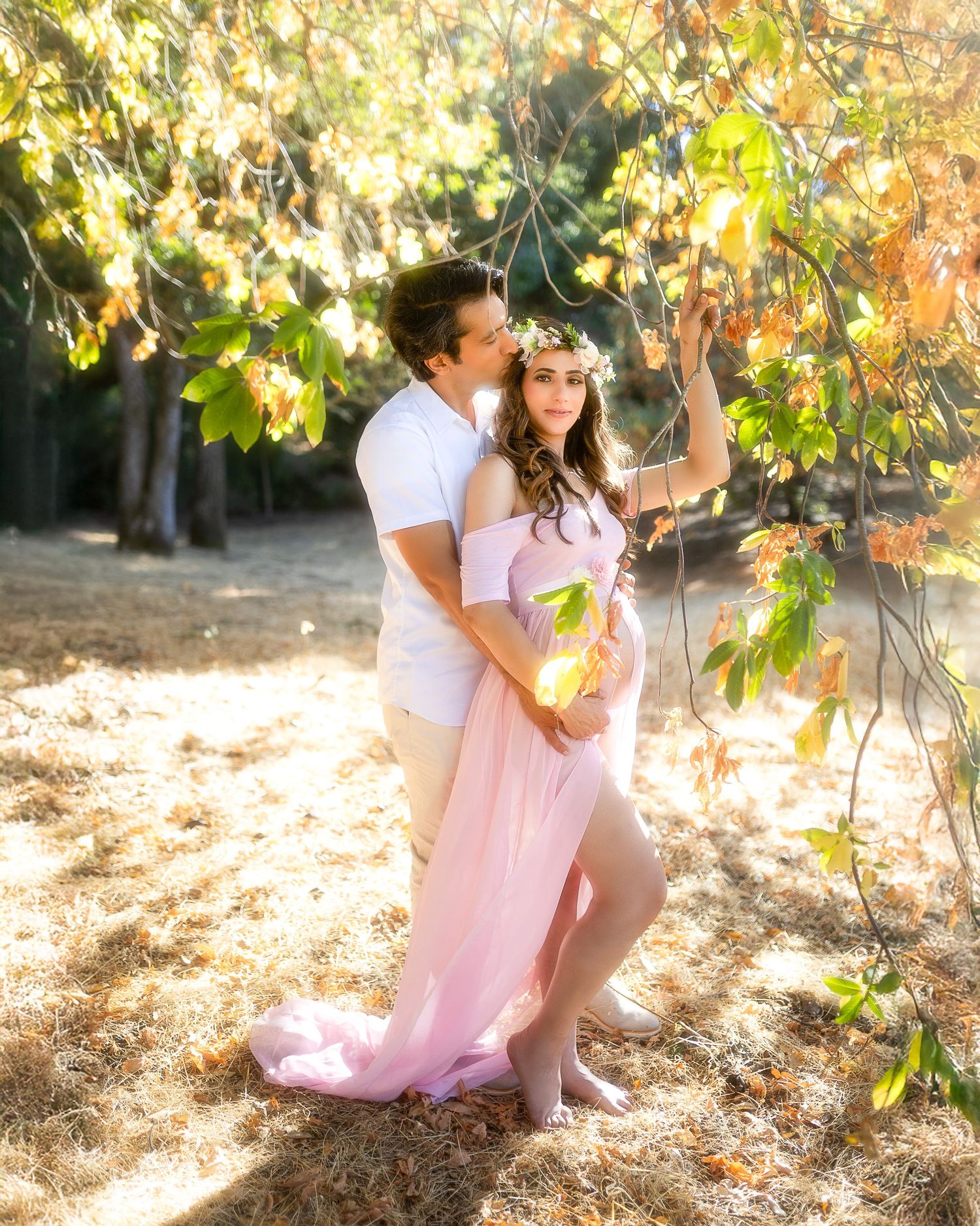 A couple poses romantically under autumn trees with sunlight filtering through the leaves.