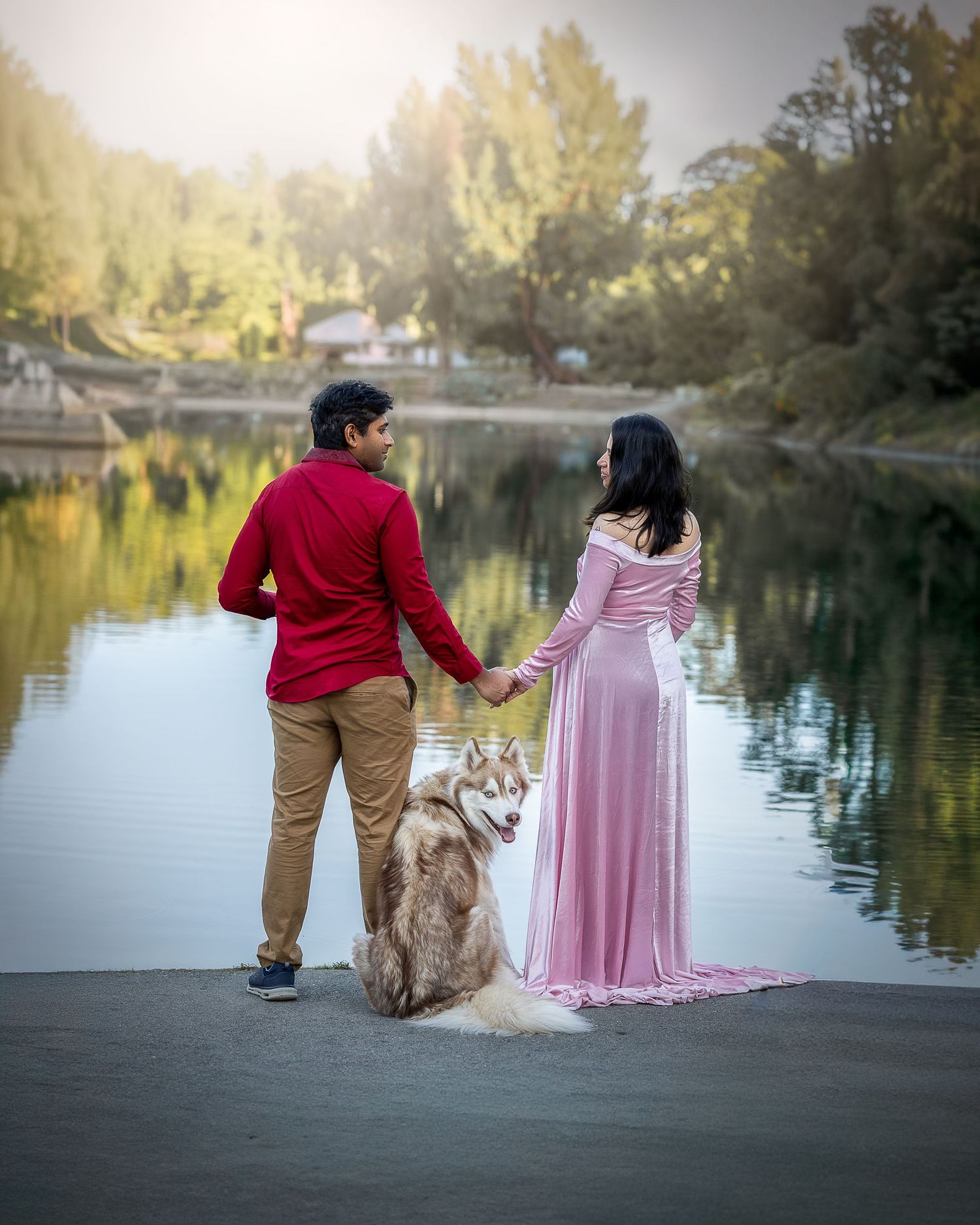 A couple in casual and formal attire stands with their husky dog by a tranquil lake during golden hour.