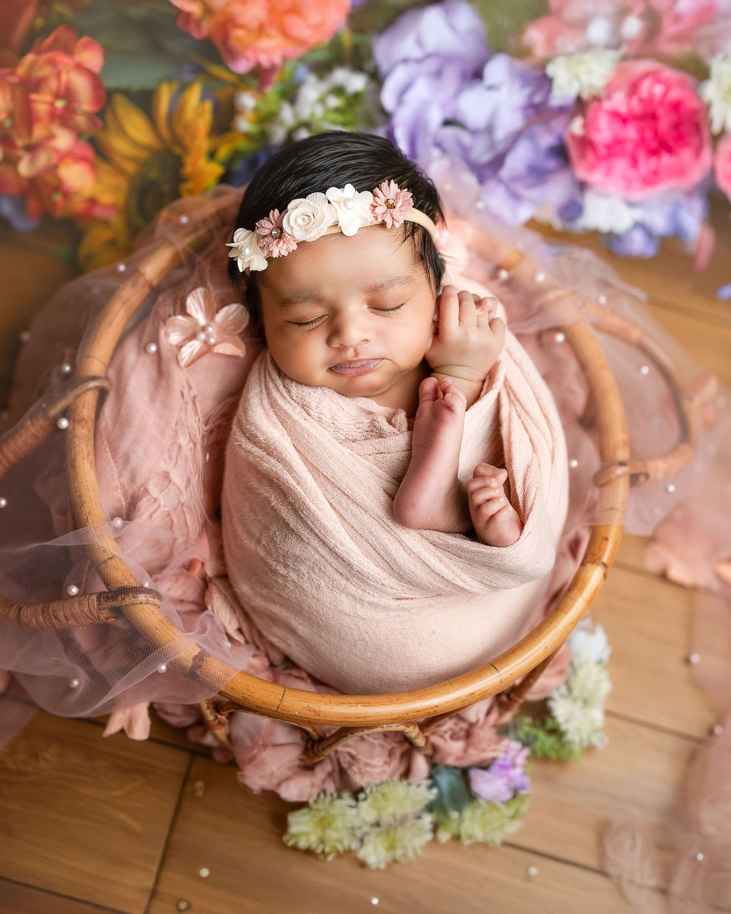 A newborn baby wrapped in pink fabric sleeps peacefully in a wooden bowl surrounded by delicate flowers.
