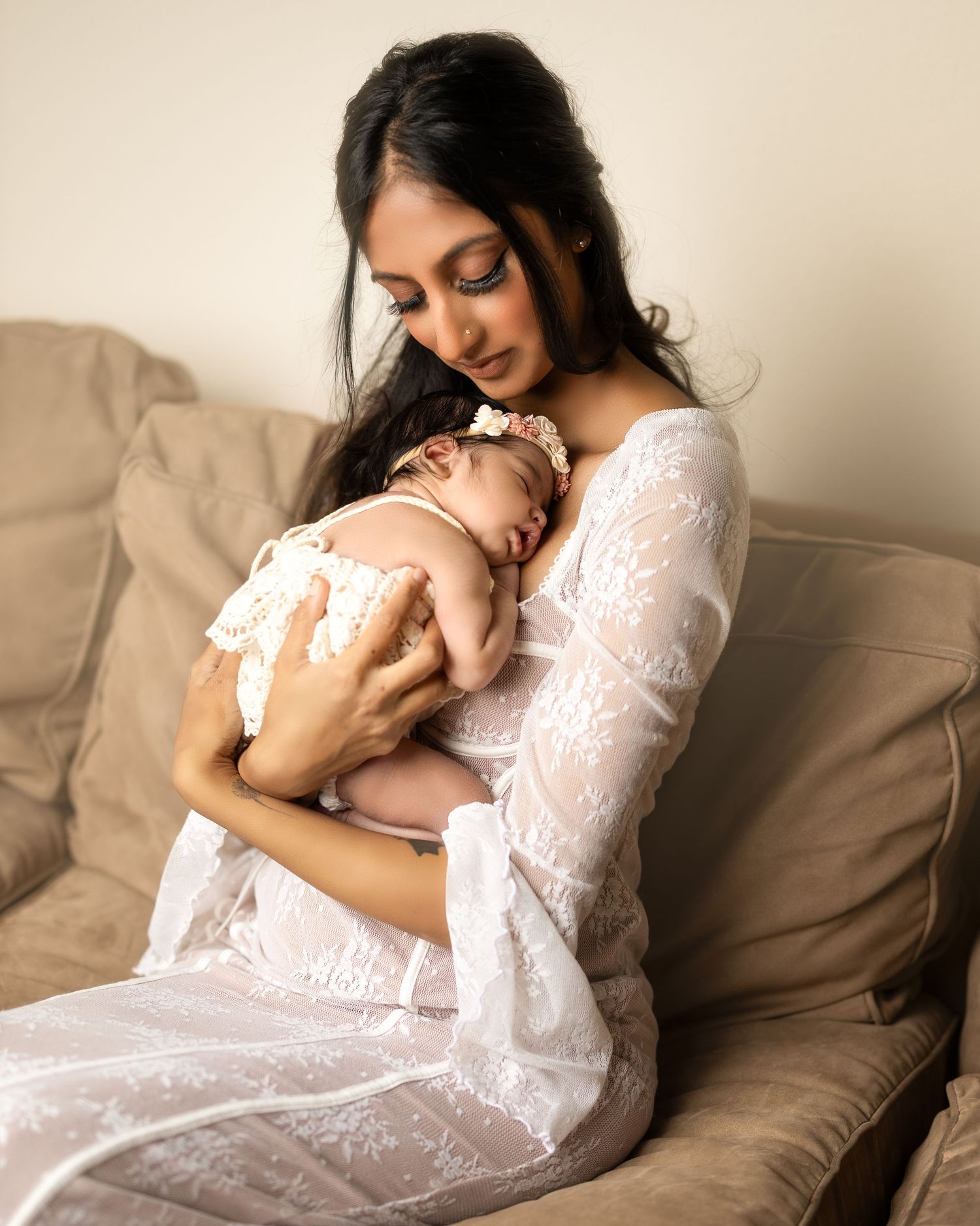 A tender moment captured as a mother in a white lace dress cradles her sleeping baby on a brown couch.