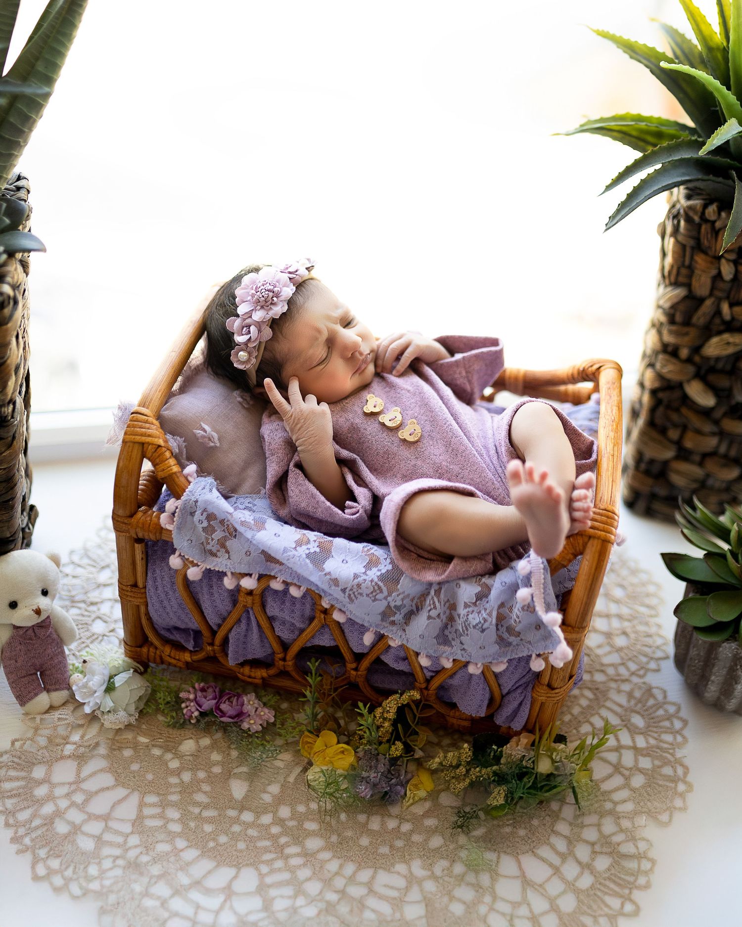 Small wicker crib decorated with lace and flowers contains a baby doll in purple outfit resting on decorative bedding.