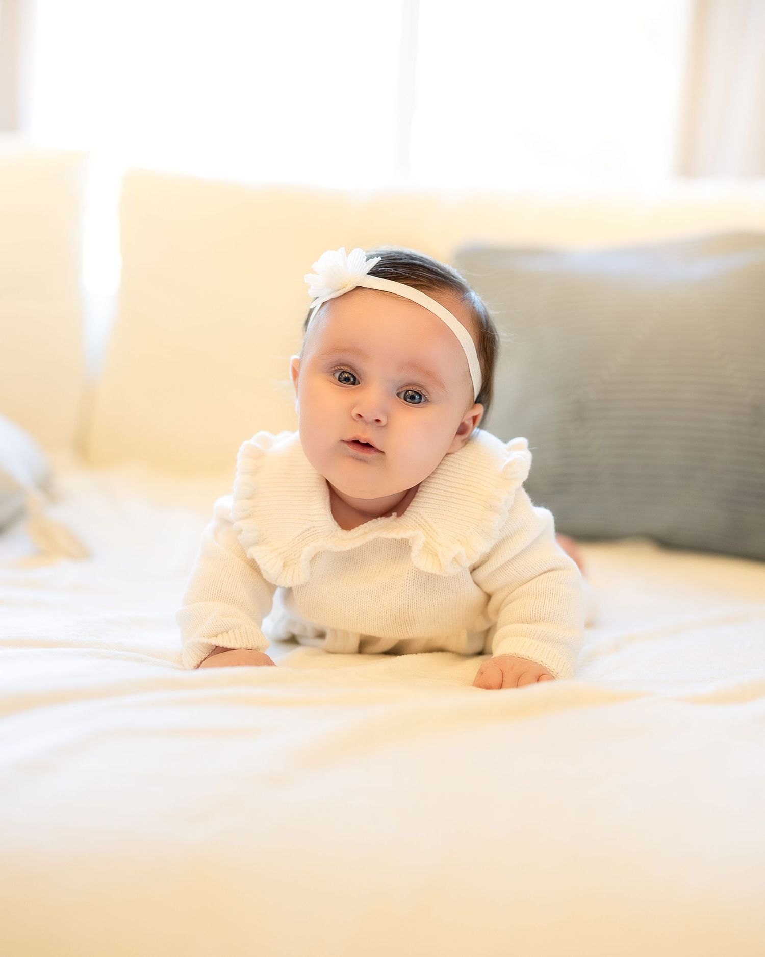 Baby in cream-colored outfit lying on soft white bedding in natural sunlit room.