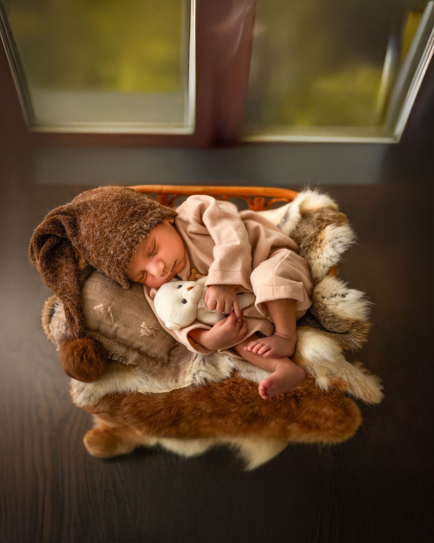 A sleeping baby nestled in cozy brown fur blankets and plush toys by a window.