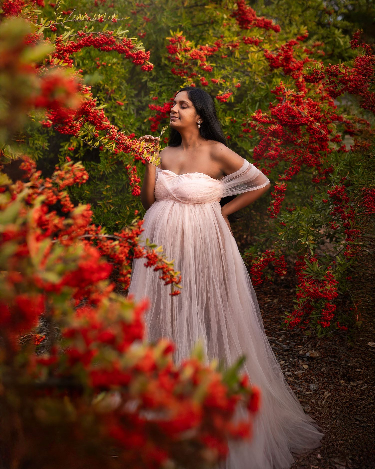 An elegant maternity portrait showcases a flowing pink dress against vibrant red flowering bushes in a garden setting.