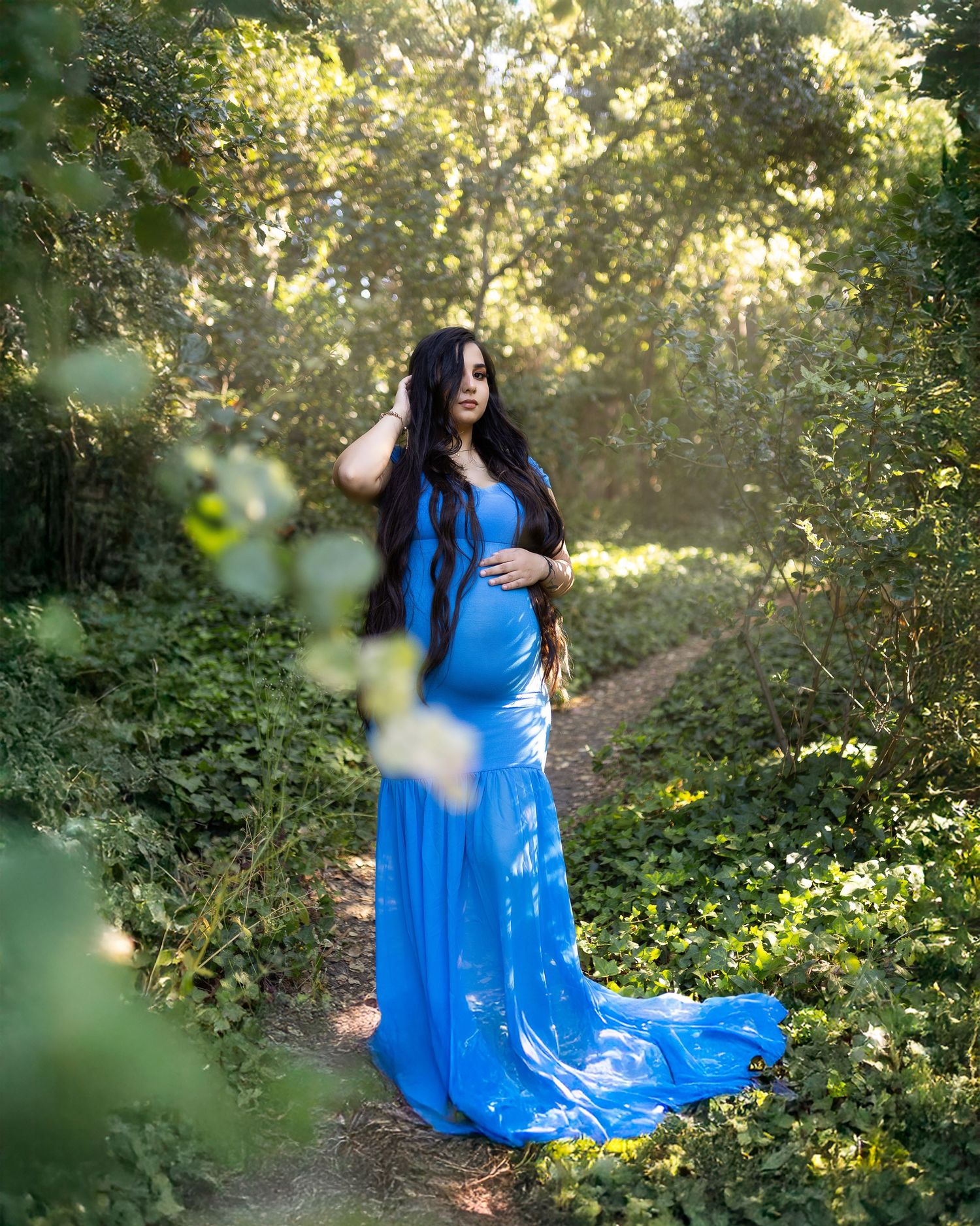 A flowing blue maternity gown catches sunlight in a dreamy forest setting with green foliage and golden light.