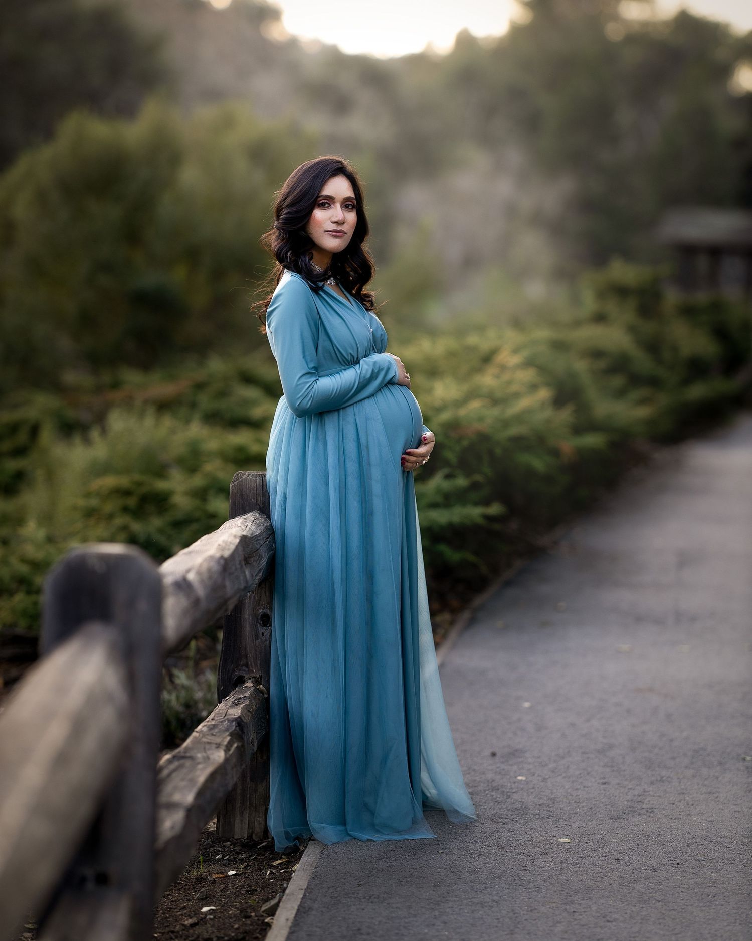 Dreamy maternity session along a park path featuring a flowing turquoise gown at sunset.