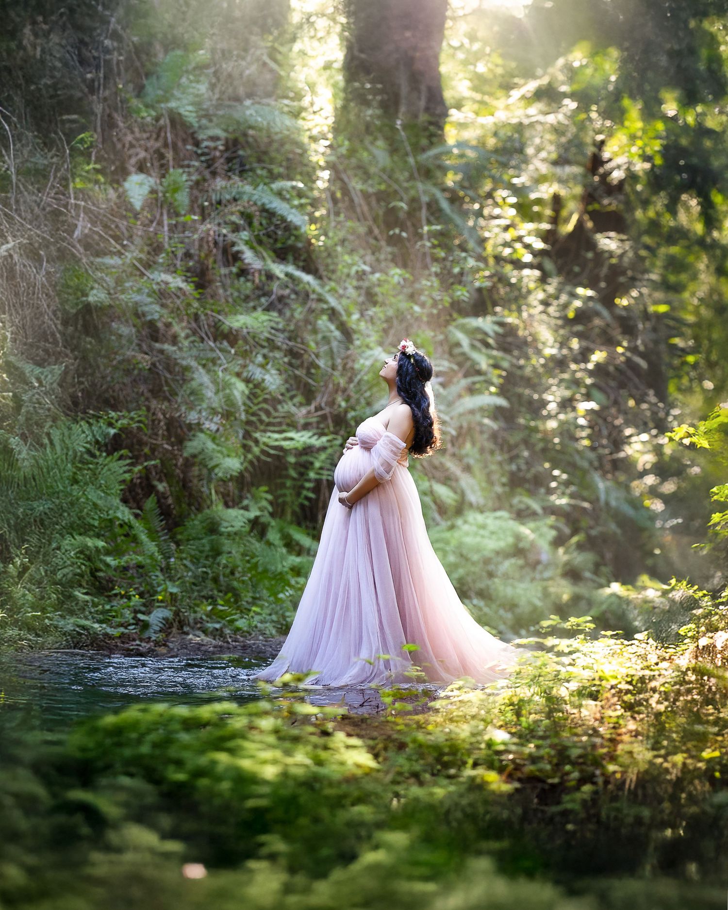 A figure in a flowing pink gown stands in a dreamy sunlit forest clearing beside a pond.