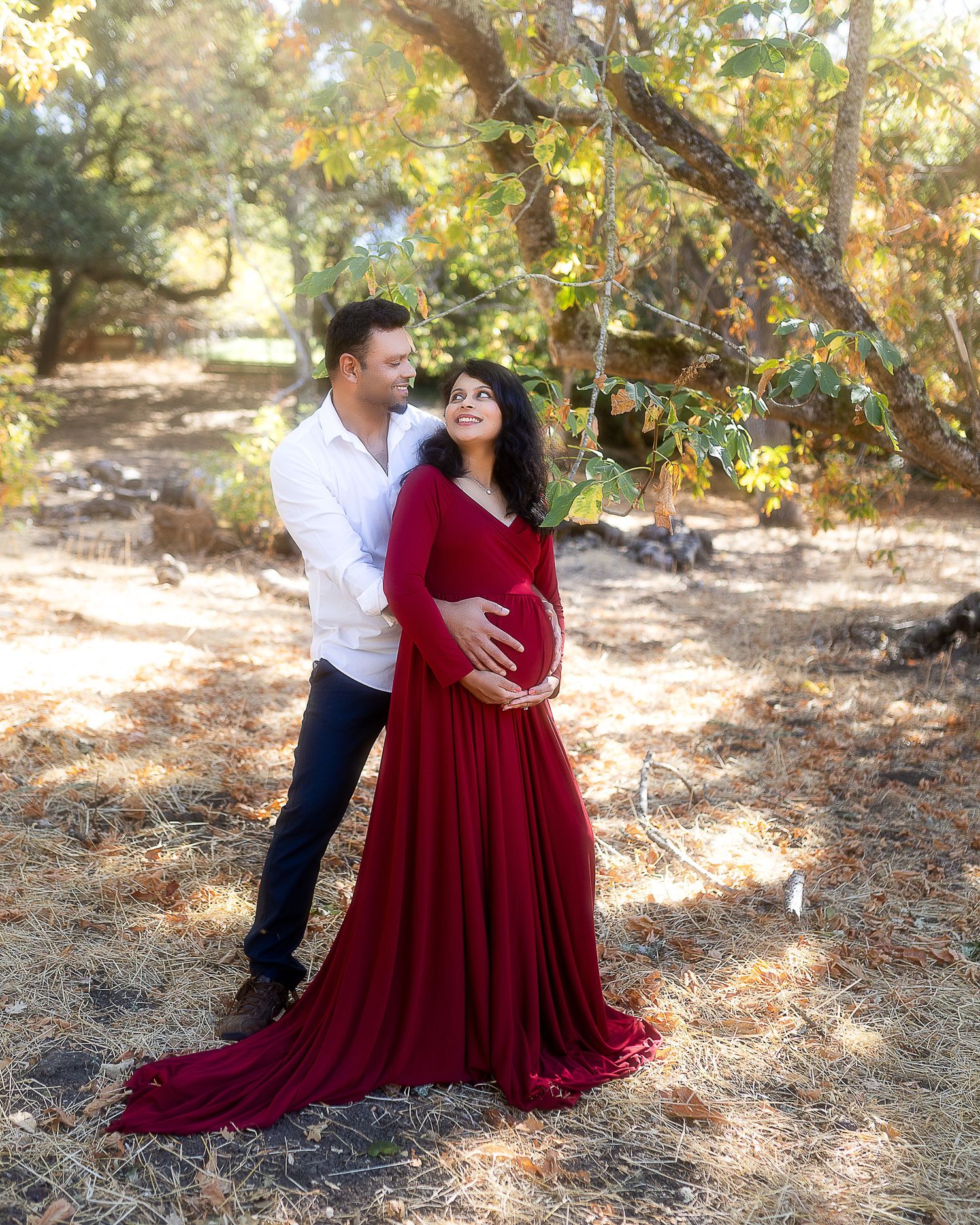 A couple embraces in a romantic outdoor photoshoot wearing burgundy dress and white shirt among autumn trees.