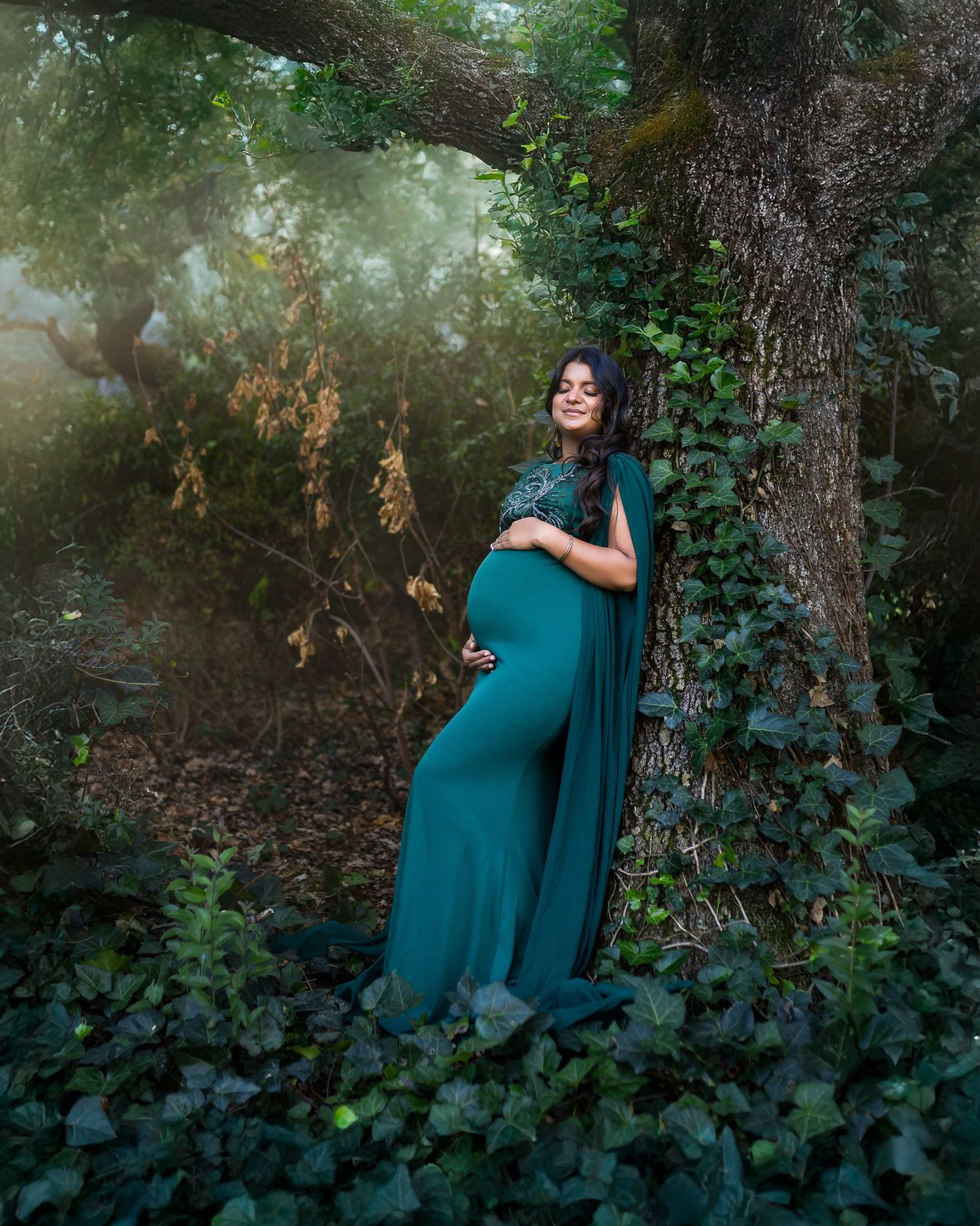 A person in a teal dress poses gracefully against a backdrop of lush greenery and trees in a moody forest setting.