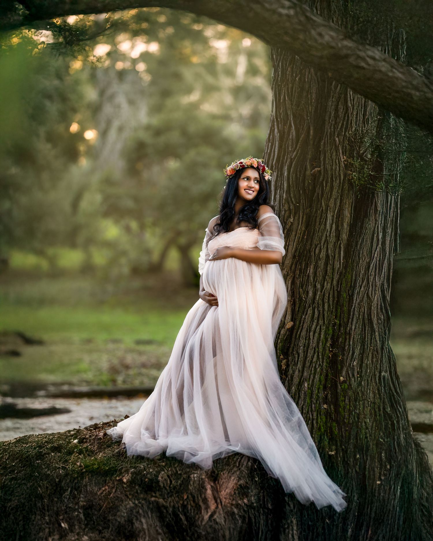 A dreamy maternity photo shows an expectant mother in a flowing white dress and floral crown leaning against a large tree.