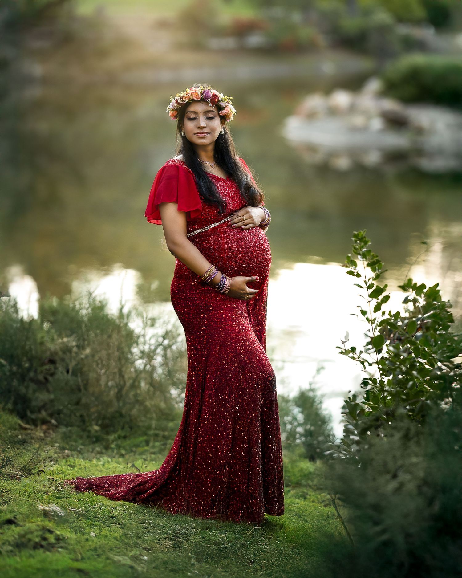 A person in a flowing burgundy polka dot dress poses gracefully by a tranquil pond in a garden setting.