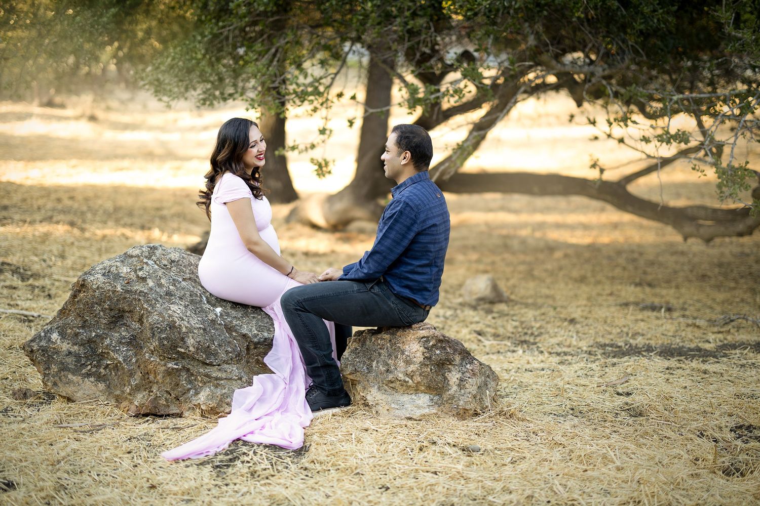 A couple sits together on a large rock in a sun-dappled outdoor setting wearing formal attire.