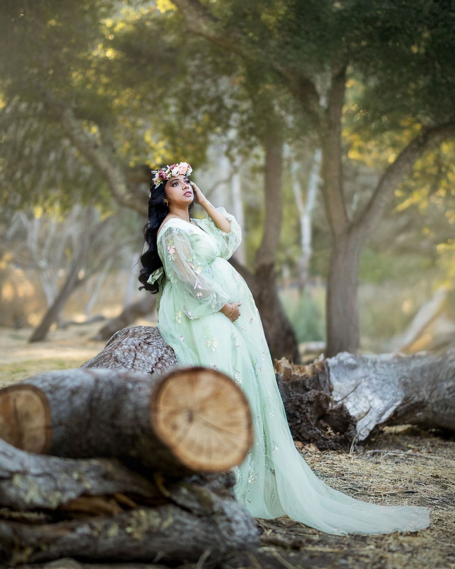A dreamy woodland scene features a person in a flowing mint green dress posing gracefully beside fallen logs.