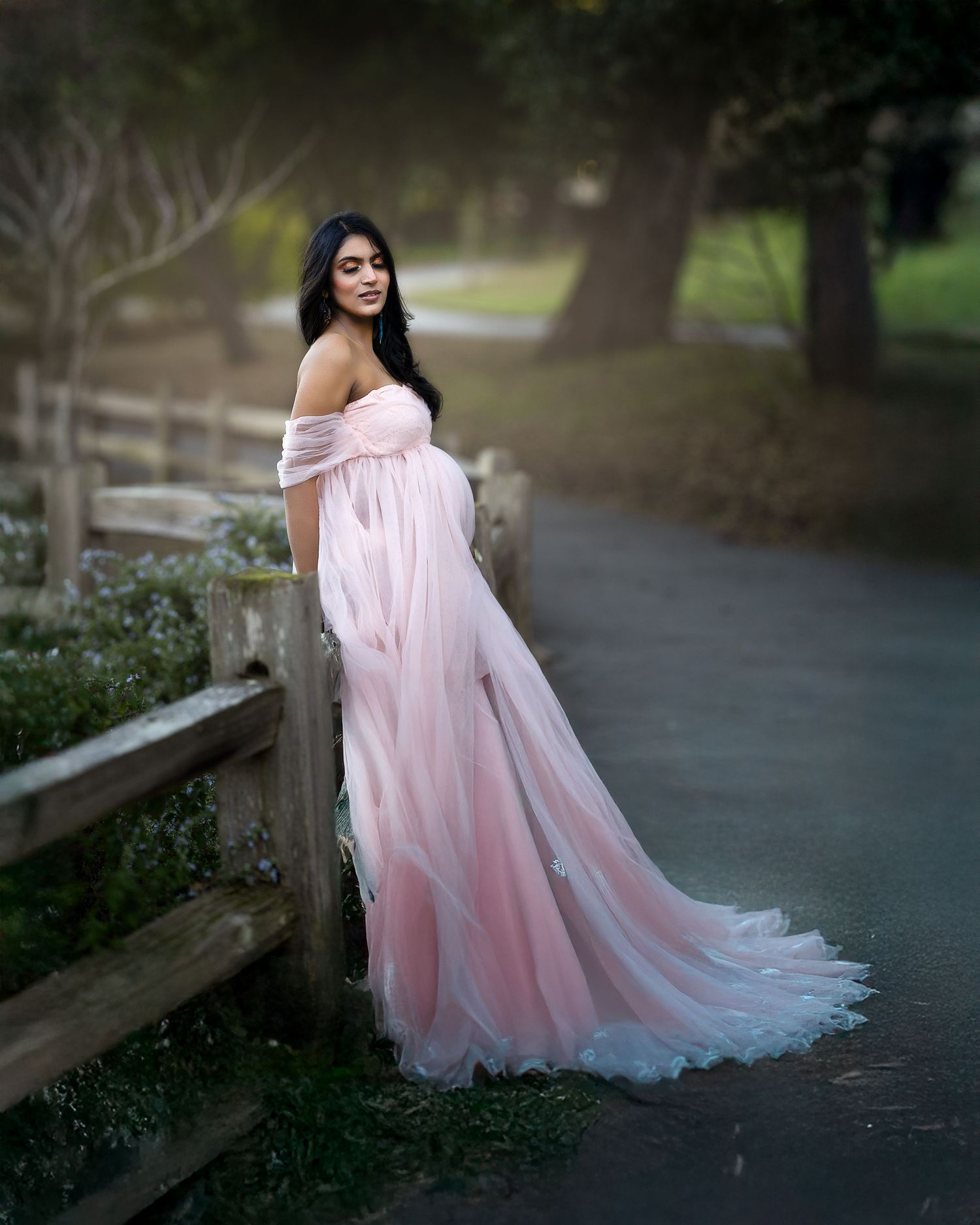 A flowing pink off-shoulder maternity gown photographed on a path with wooden fencing and trees in the background.